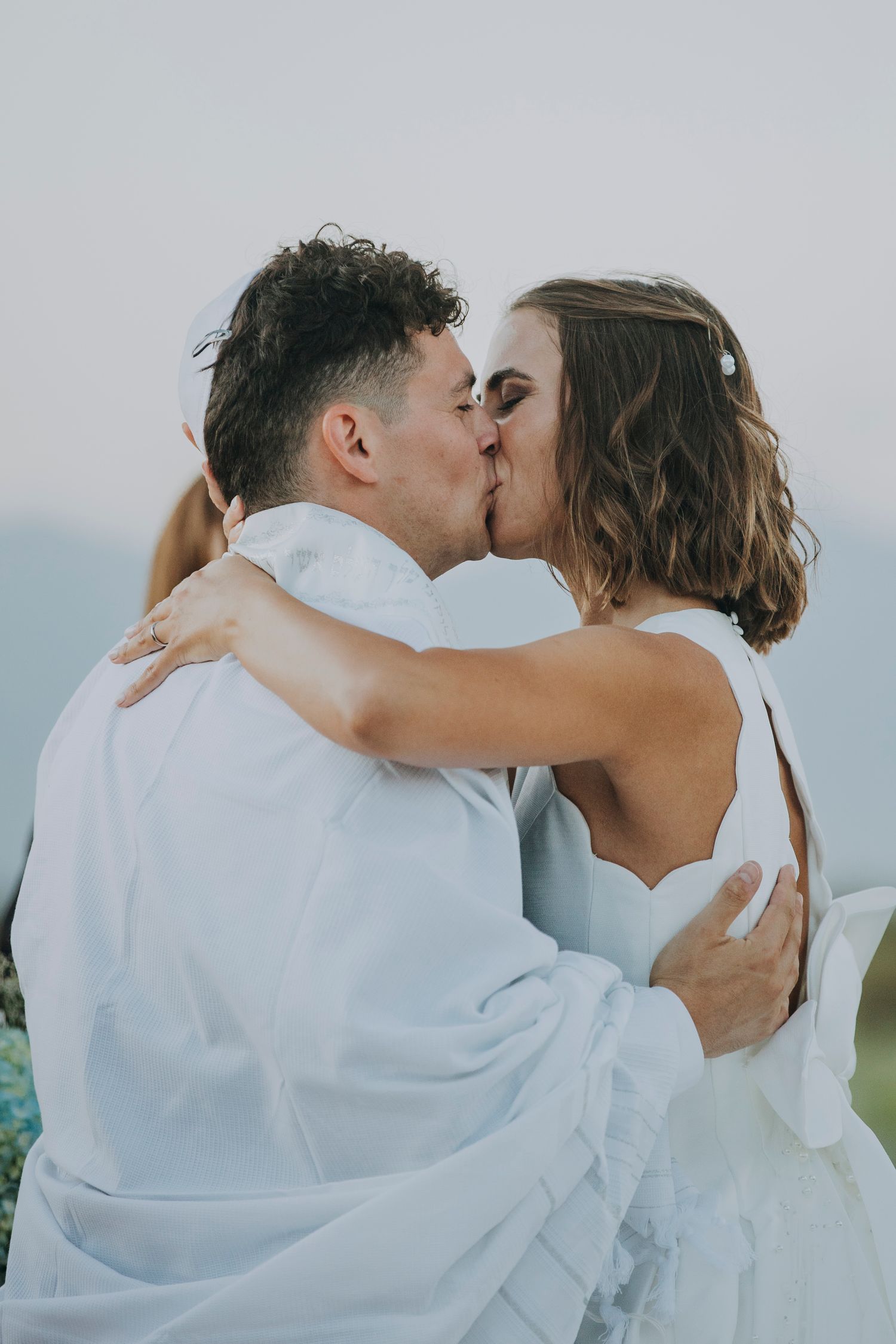 Newlywed couple shares romantic first kiss at outdoor wedding ceremony against soft sky backdrop.