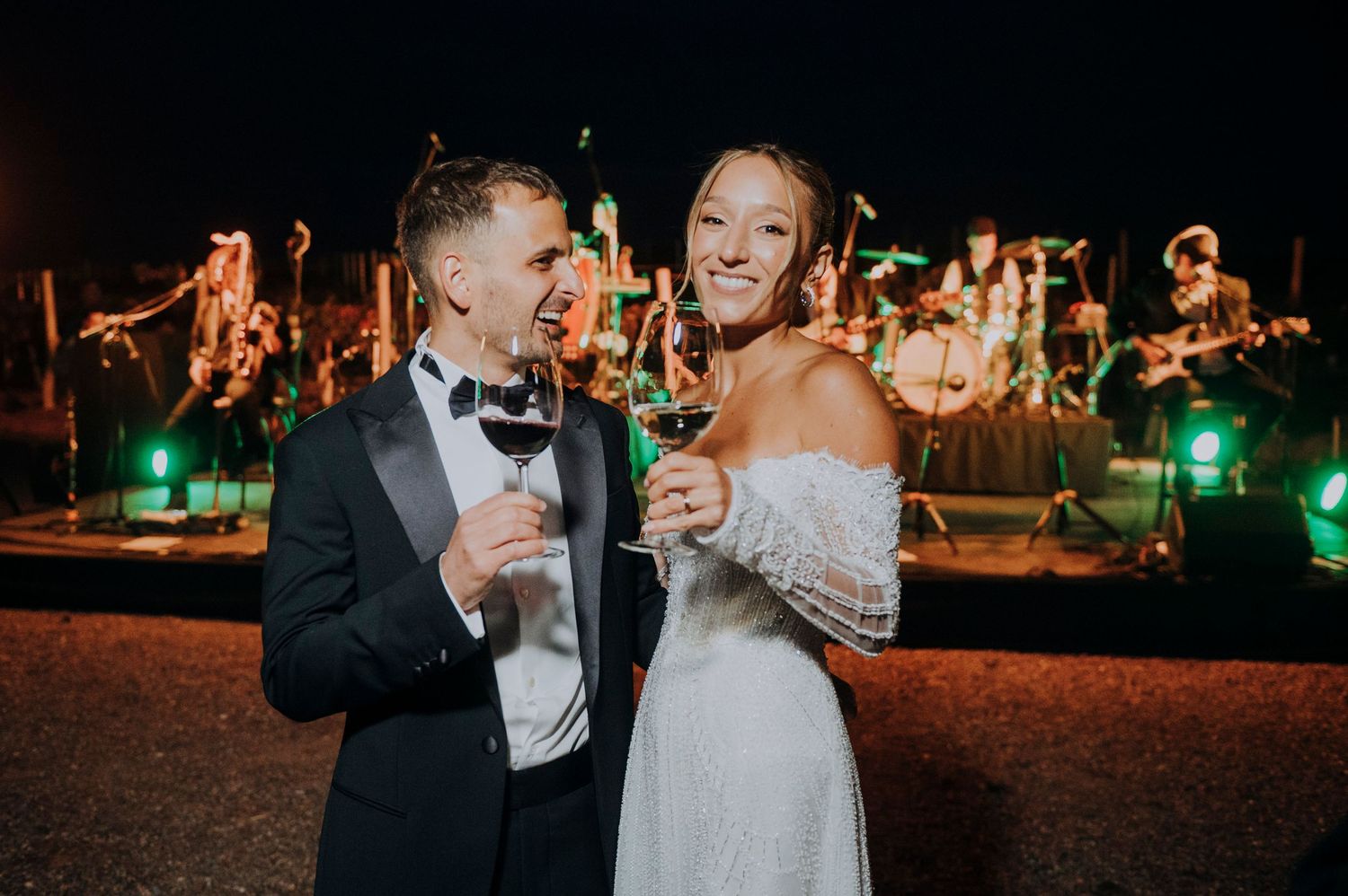 Bride and groom toasting champagne at outdoor wedding reception with live band performing on stage at night.