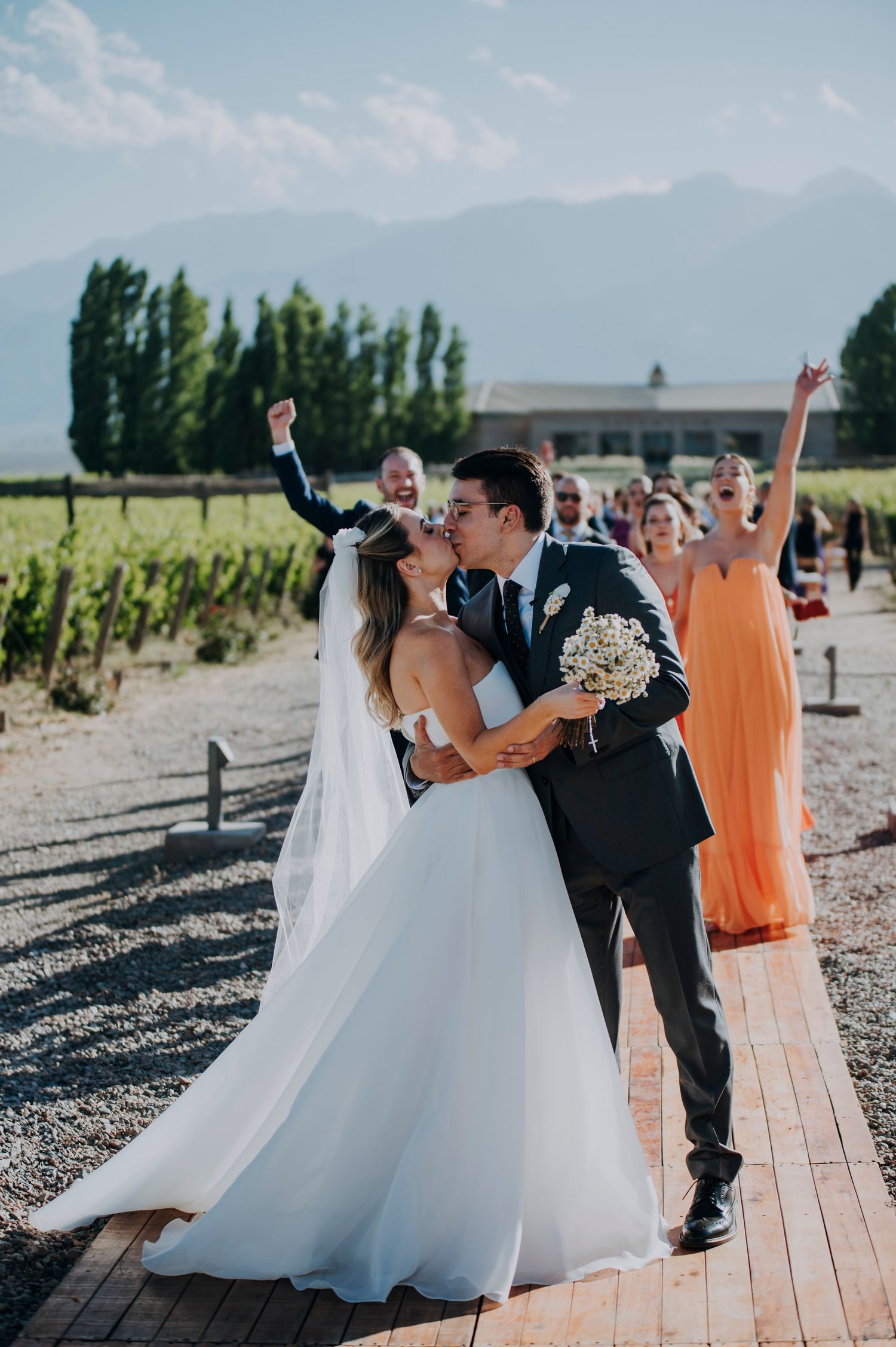 Bride and groom share a kiss while wedding party cheers behind them at a vineyard wedding venue.