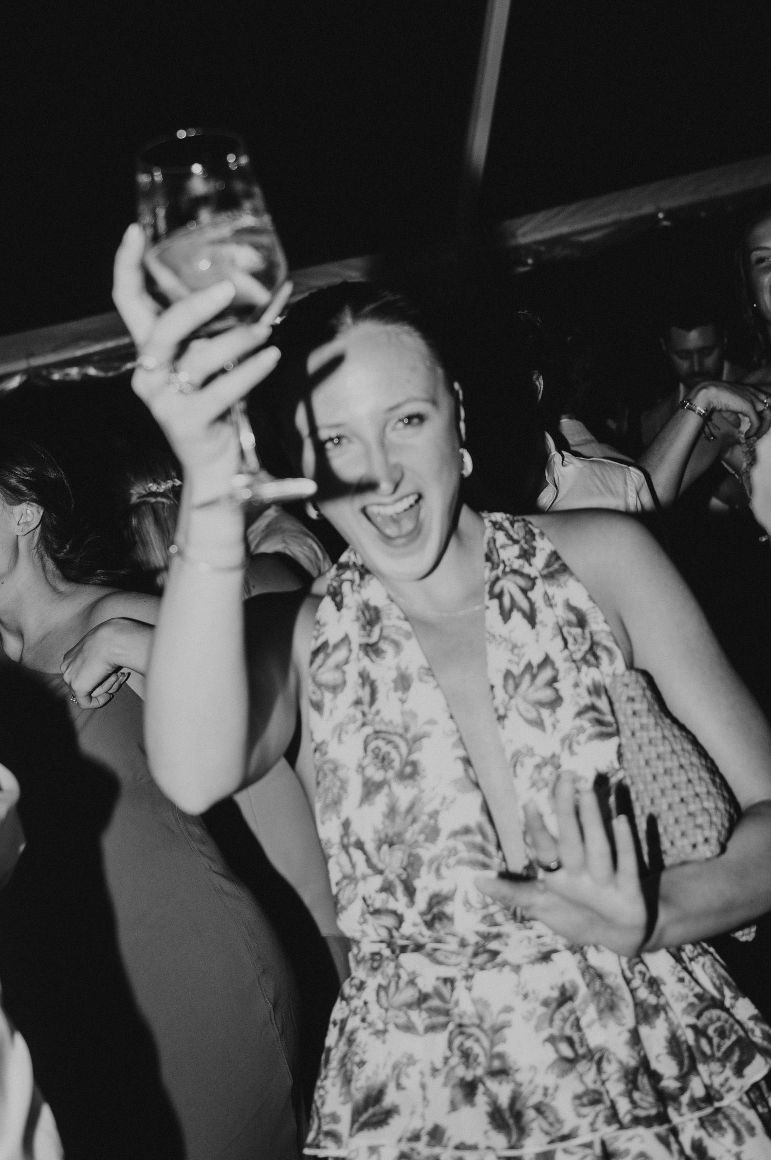 Excited woman in floral dress raising a drink glass at a lively party celebration.