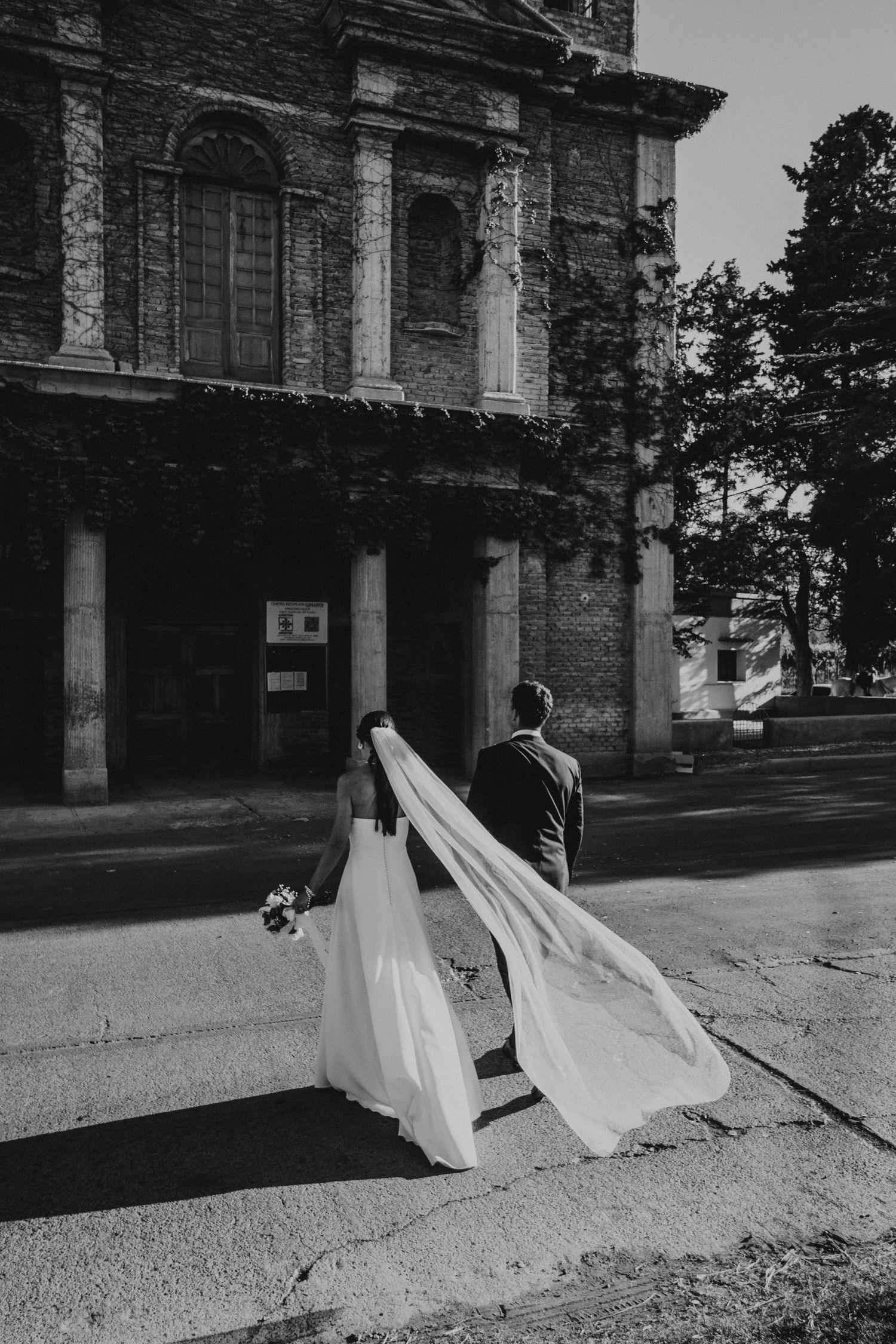 Black and white photo of bride and groom walking past historic church, veil flowing in wind. Couple portrait at sunset in Mendoza.