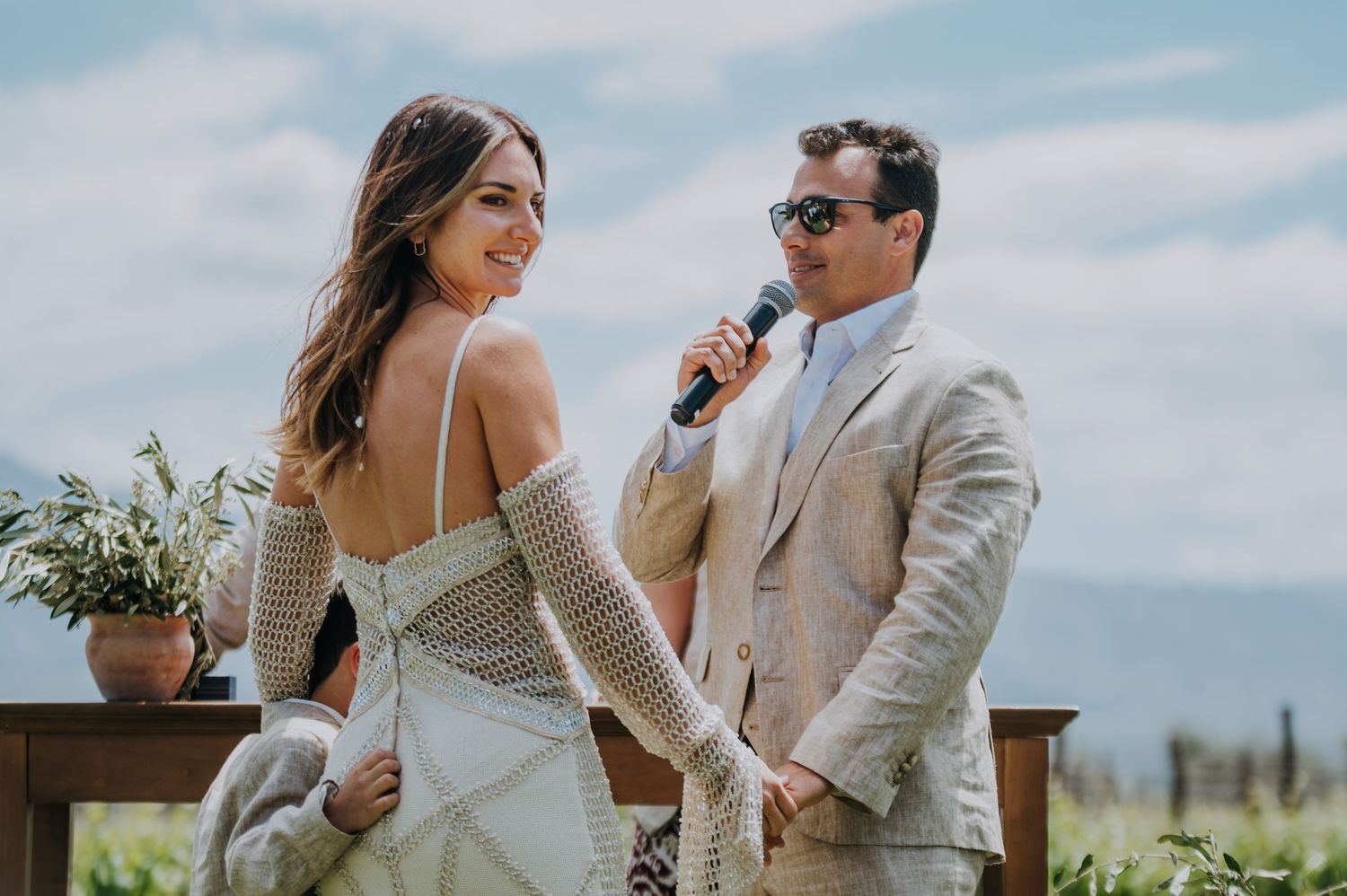 Bride and groom share a romantic moment at outdoor wedding ceremony, man in beige suit speaks into microphone.