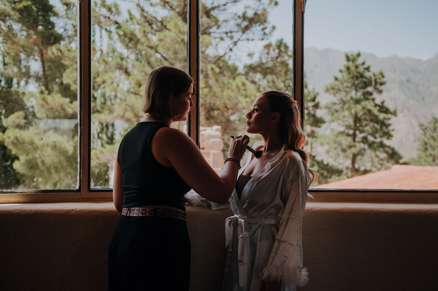 Bridesmaid helping bride with jewelry near large windows with mountain views on wedding day.