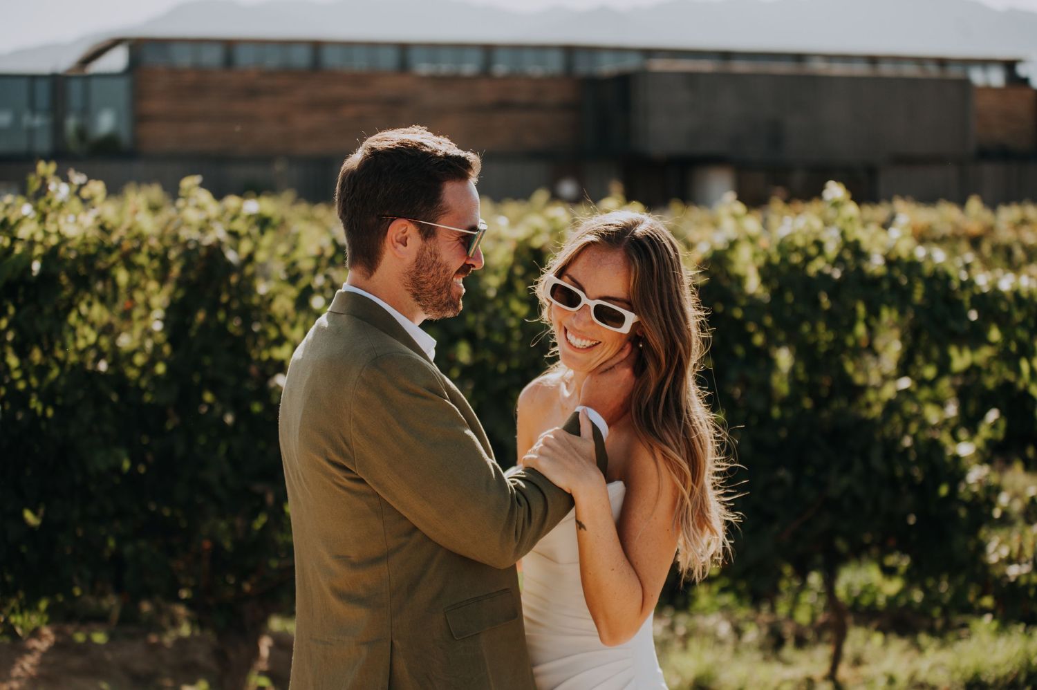 Couple smiling in vineyard, bride in white dress with sunglasses, groom in olive suit, golden hour lighting.