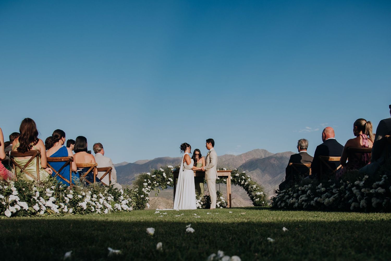 Outdoor wedding ceremony on green lawn with mountain backdrop, couple at floral arch, guests seated under clear blue sky.