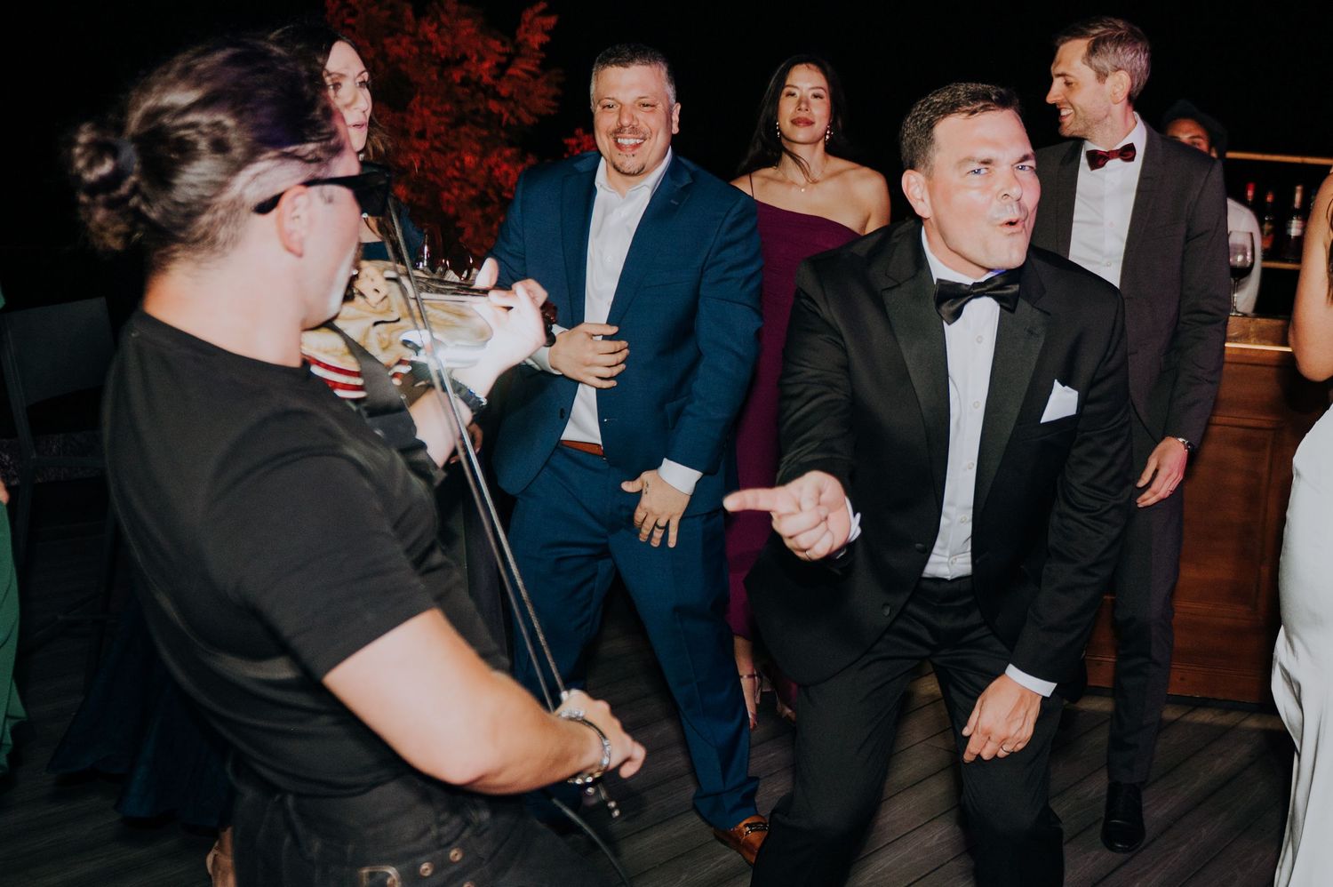 Guests dance energetically at a wedding reception while a violinist performs nearby.