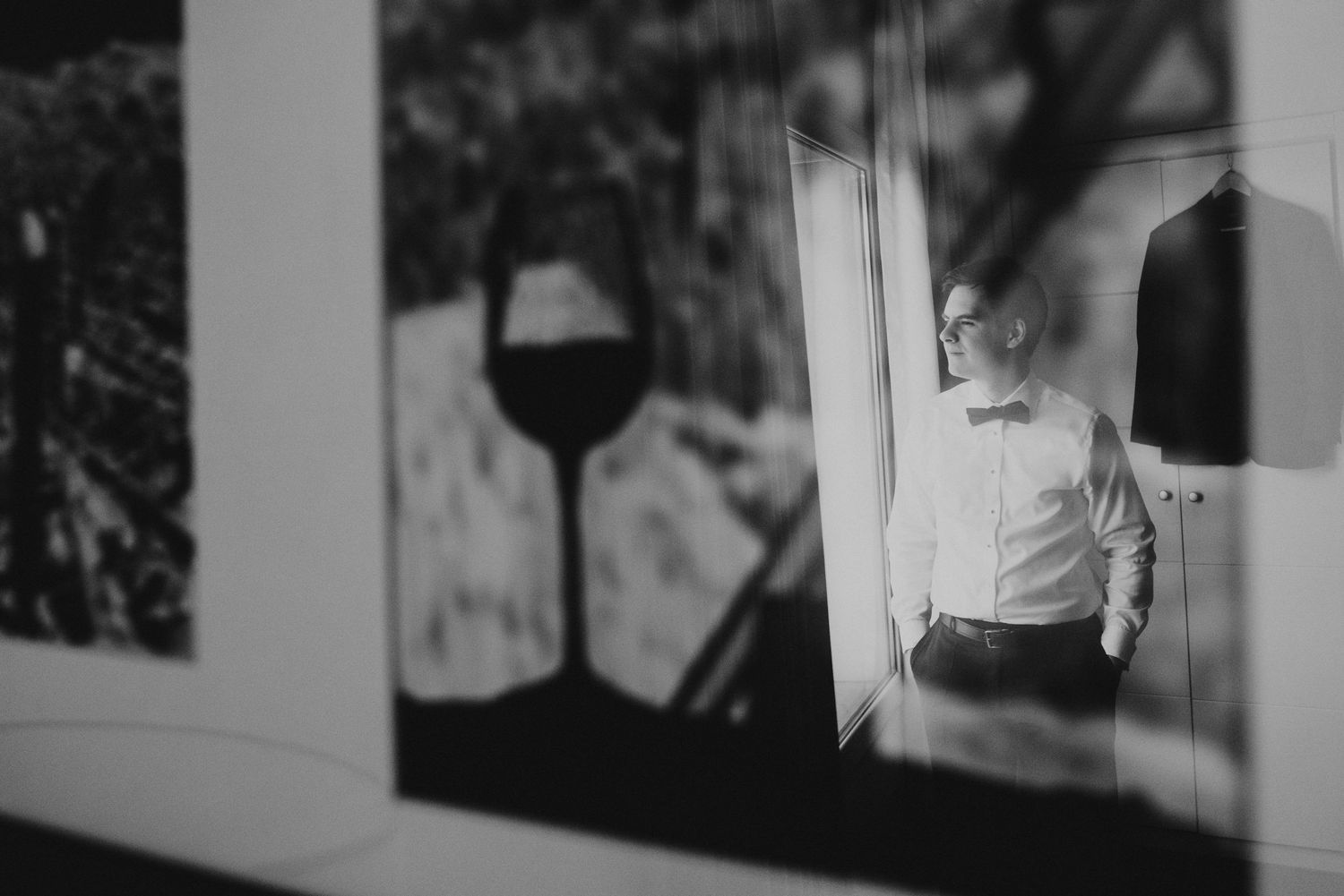 Black and white photo of a groom in bow tie and suspenders standing by a window on his wedding day.