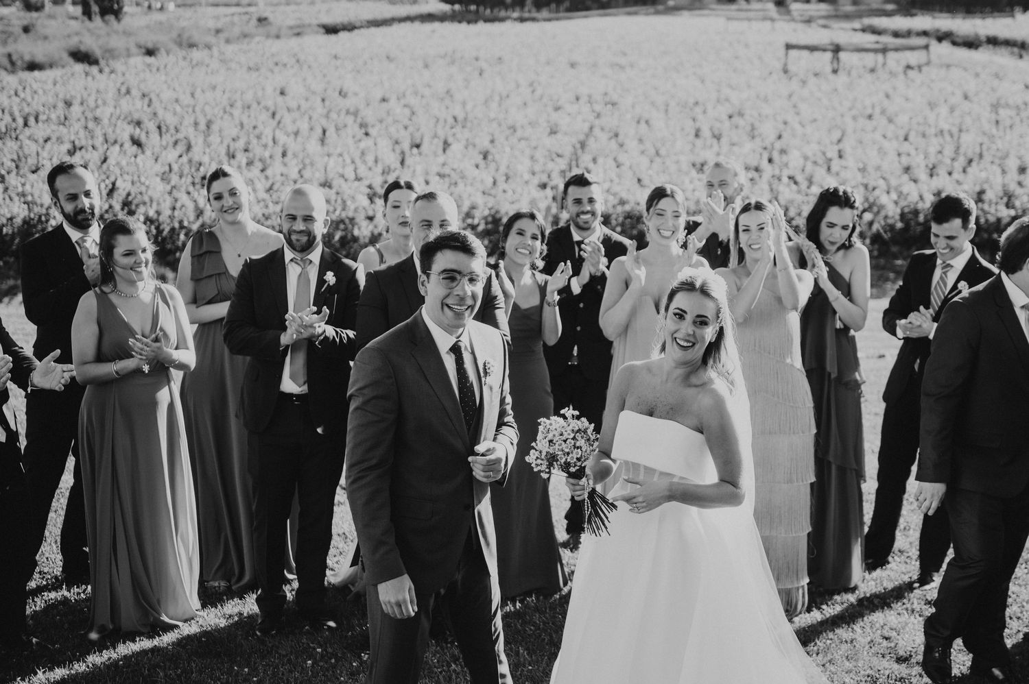 Black and white photo of smiling bride and groom with wedding party celebrating in vineyard.