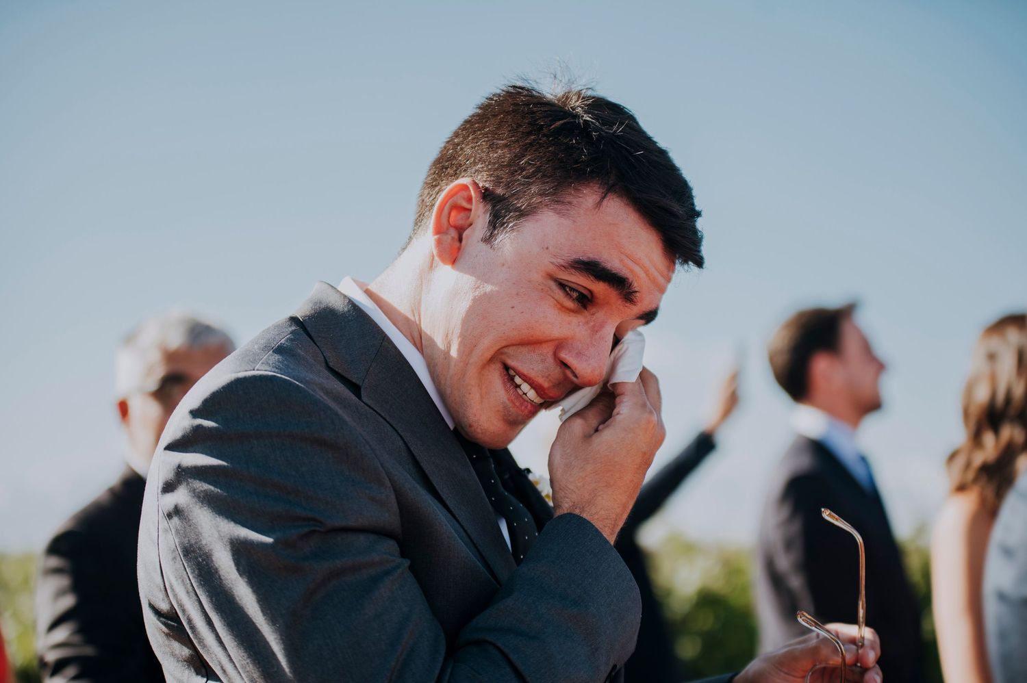 Emotional groom wiping tears at outdoor wedding ceremony wearing gray suit on sunny day.