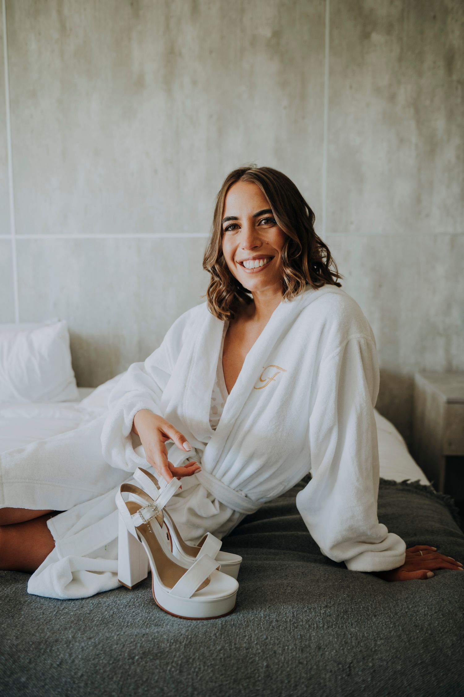 Smiling bride in white robe sits on bed holding wedding shoes during bridal preparations.