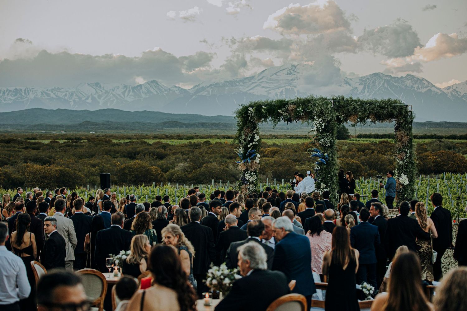 Outdoor wedding ceremony with floral arch, guests gathered, stunning mountain backdrop at sunset.