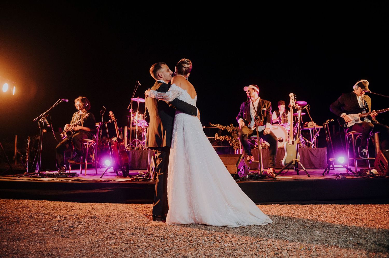 Bride and groom share first dance outdoors at night with live band performing on stage behind them.