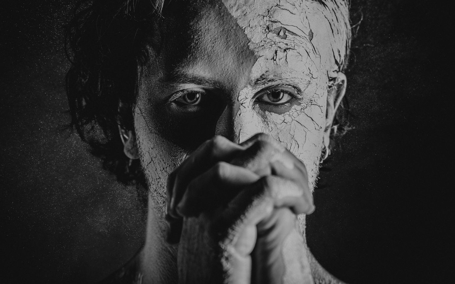 Black and white dramatic portrait of person with cracked clay face holding hands in prayer position.