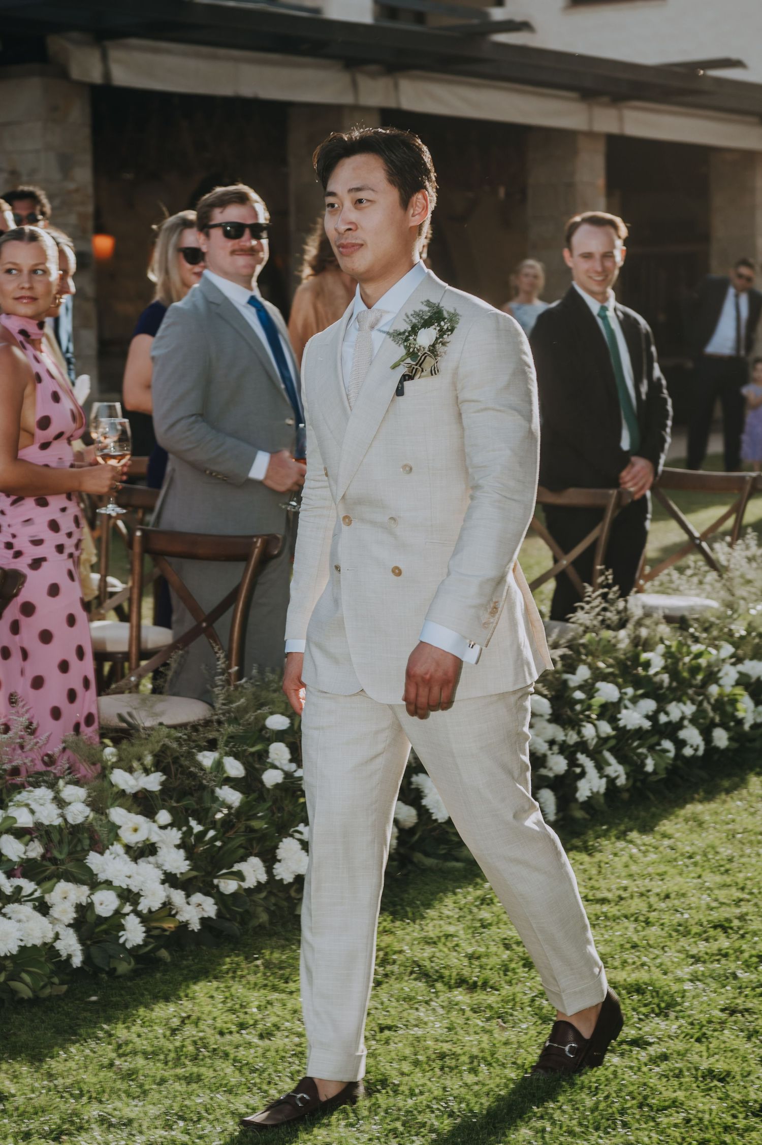 Groom walks confidently in cream double-breasted suit with floral boutonniere at outdoor wedding ceremony.