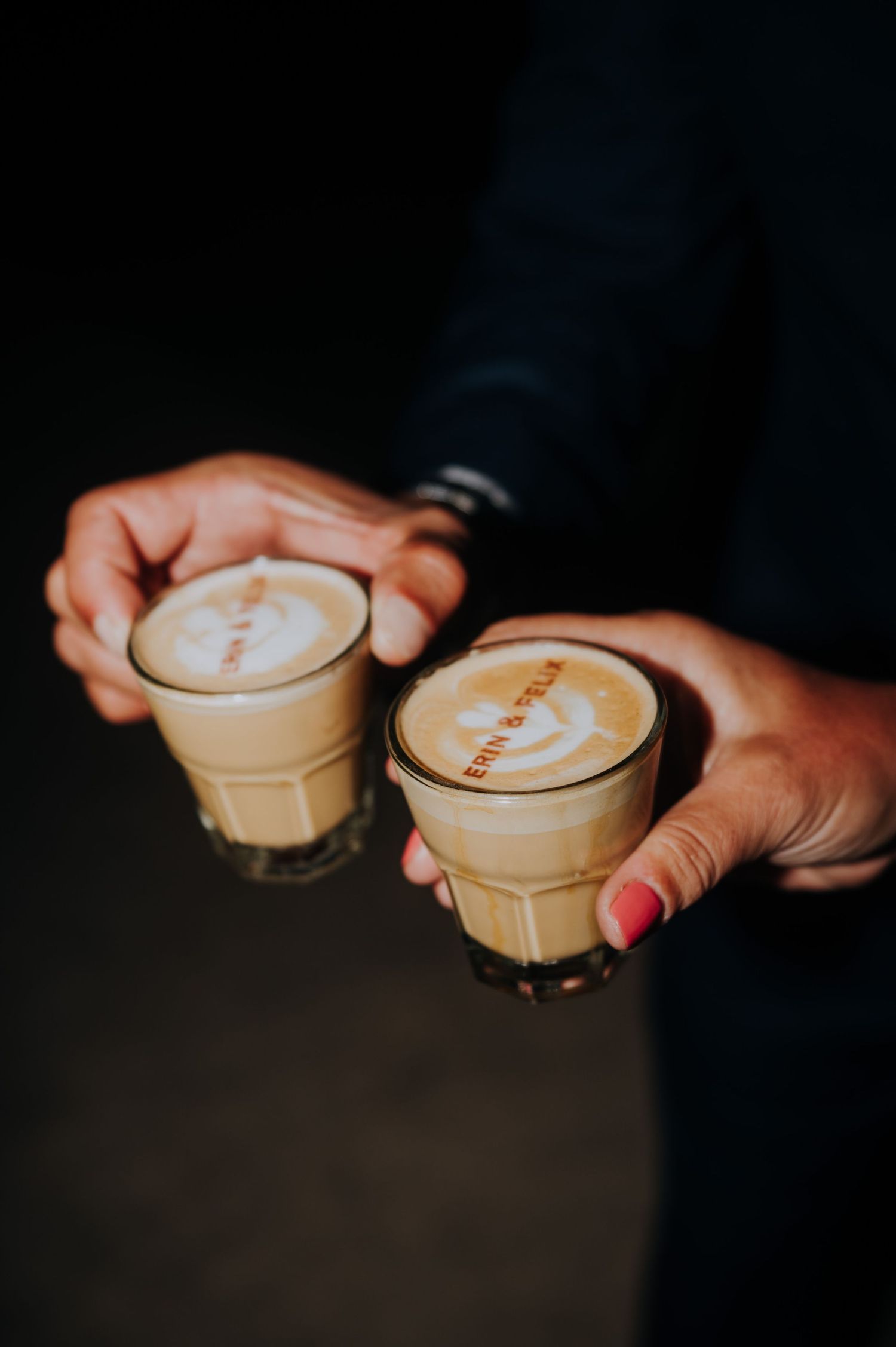 Two hands holding glass cups of latte art coffee with creamy foam tops in a dark setting.