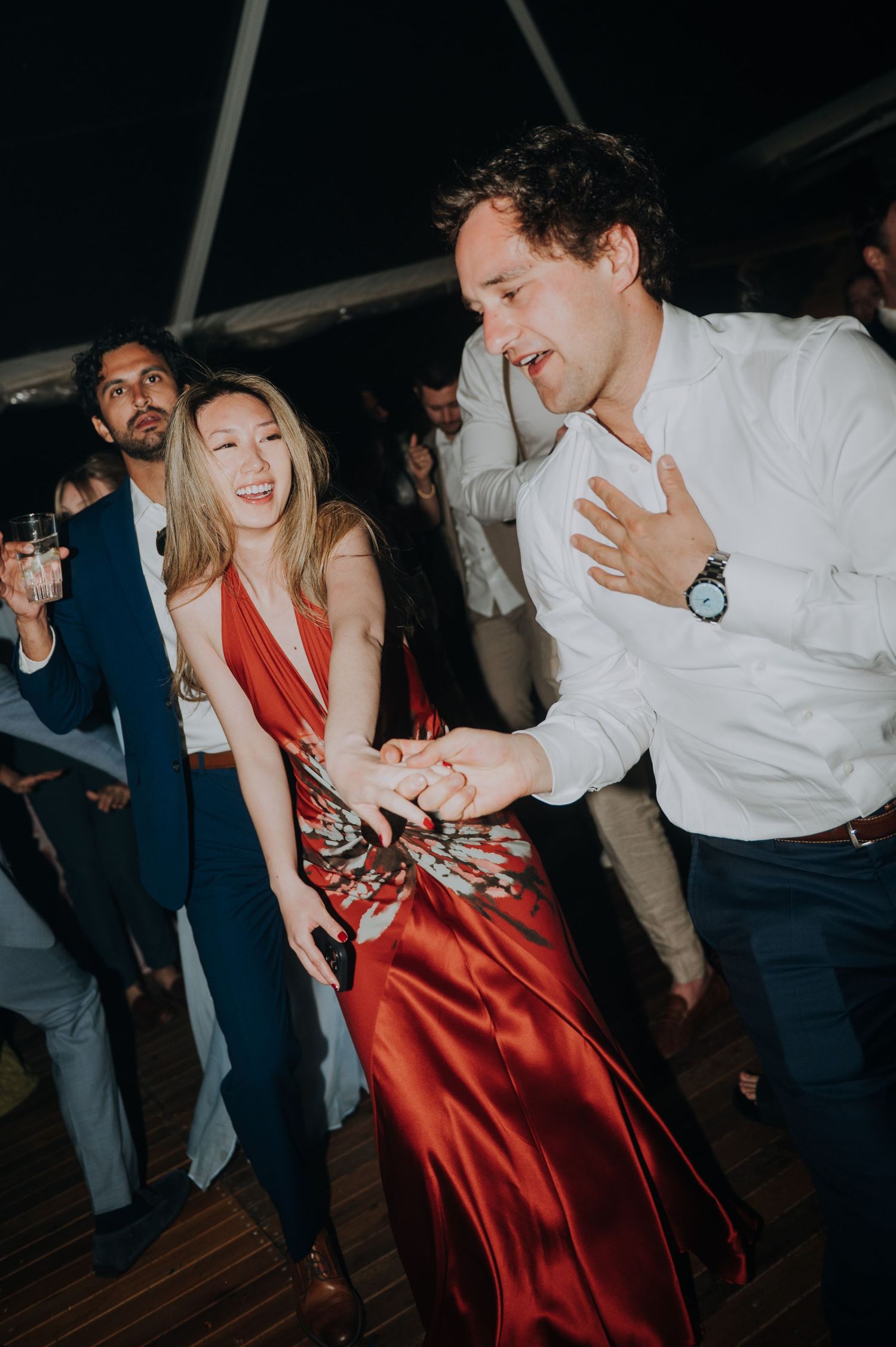 Guests dancing energetically in red satin dress and white shirt at wedding reception.