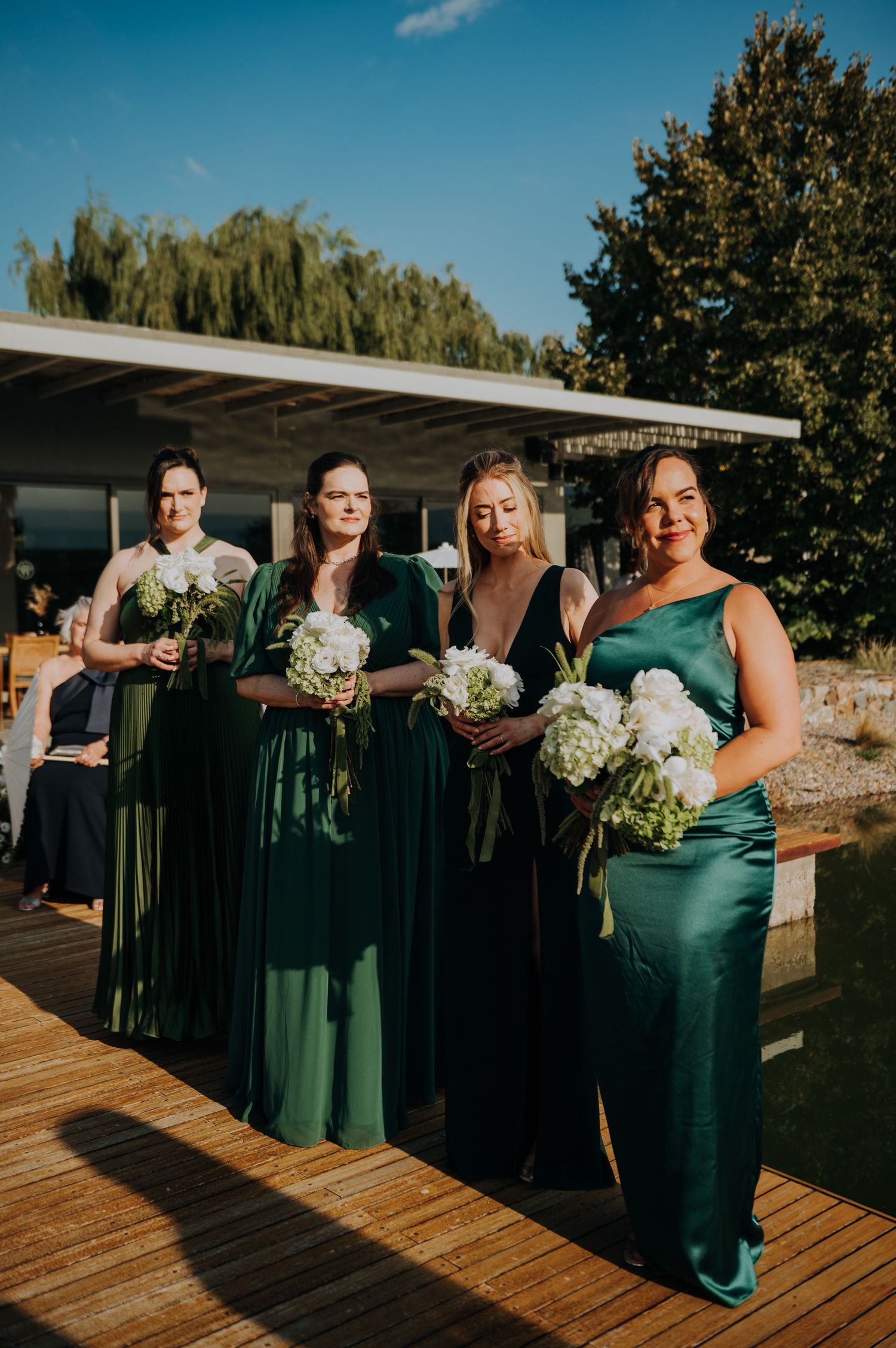 Four bridesmaids in emerald green gowns holding white hydrangea bouquets at outdoor waterfront wedding ceremony.
