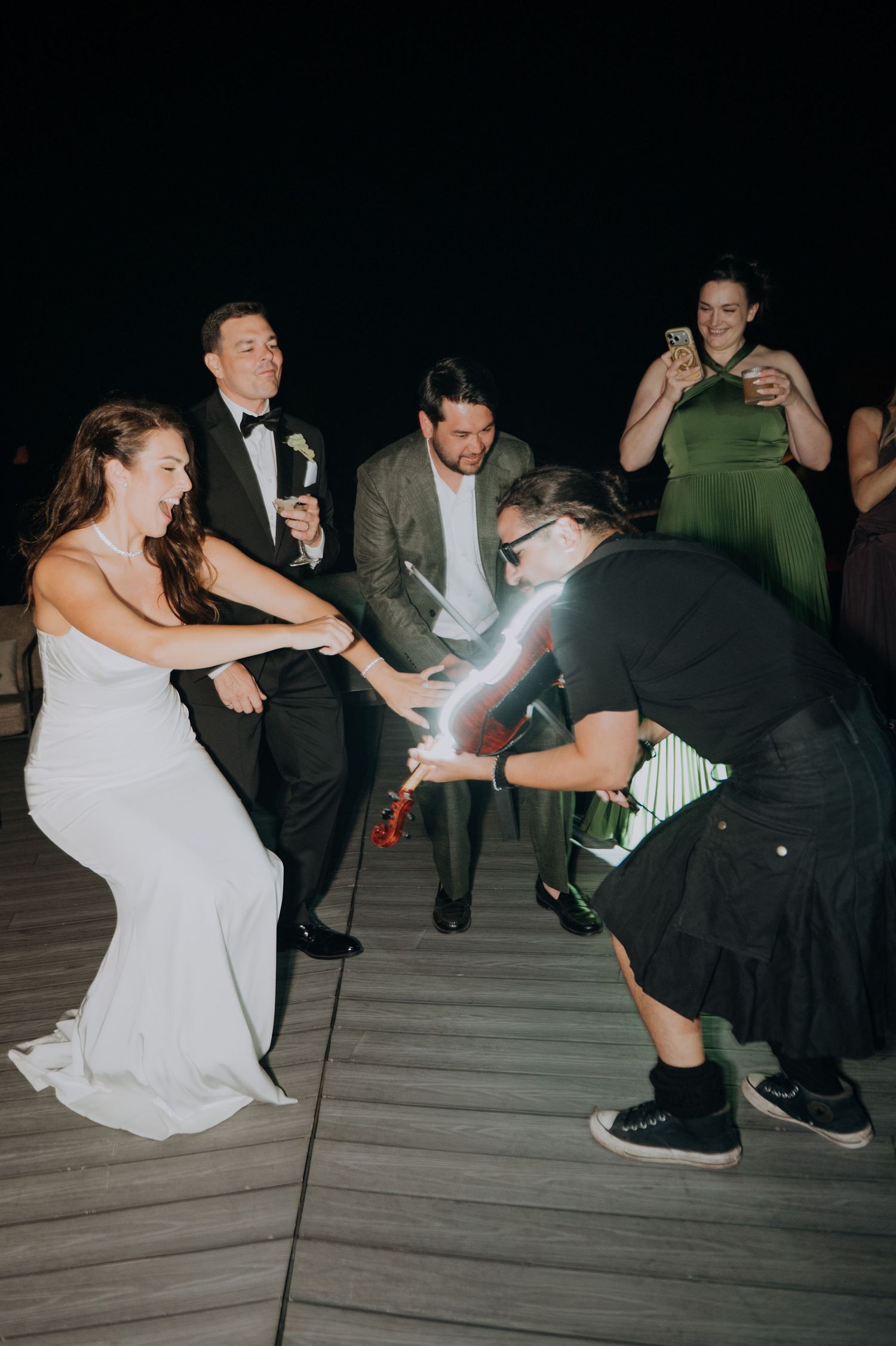 Bride and groom laugh as guest pops champagne bottle with sparkler at lively outdoor wedding reception at night.