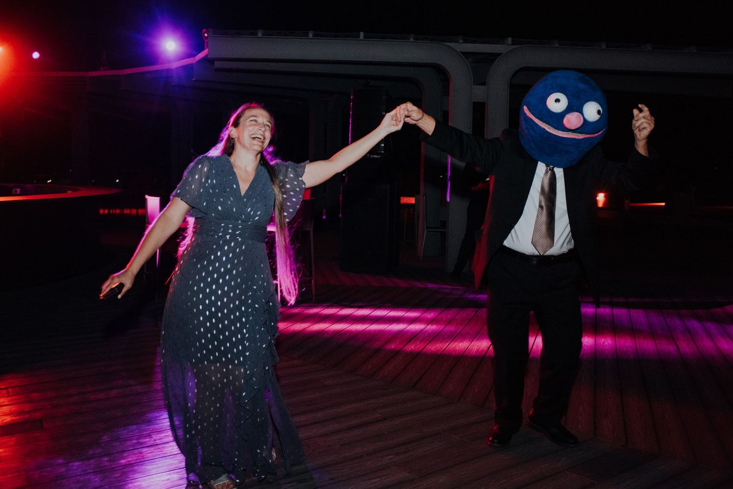 Wedding guest in blue monster costume dancing with woman in sparkly dress on dimly lit dance floor.