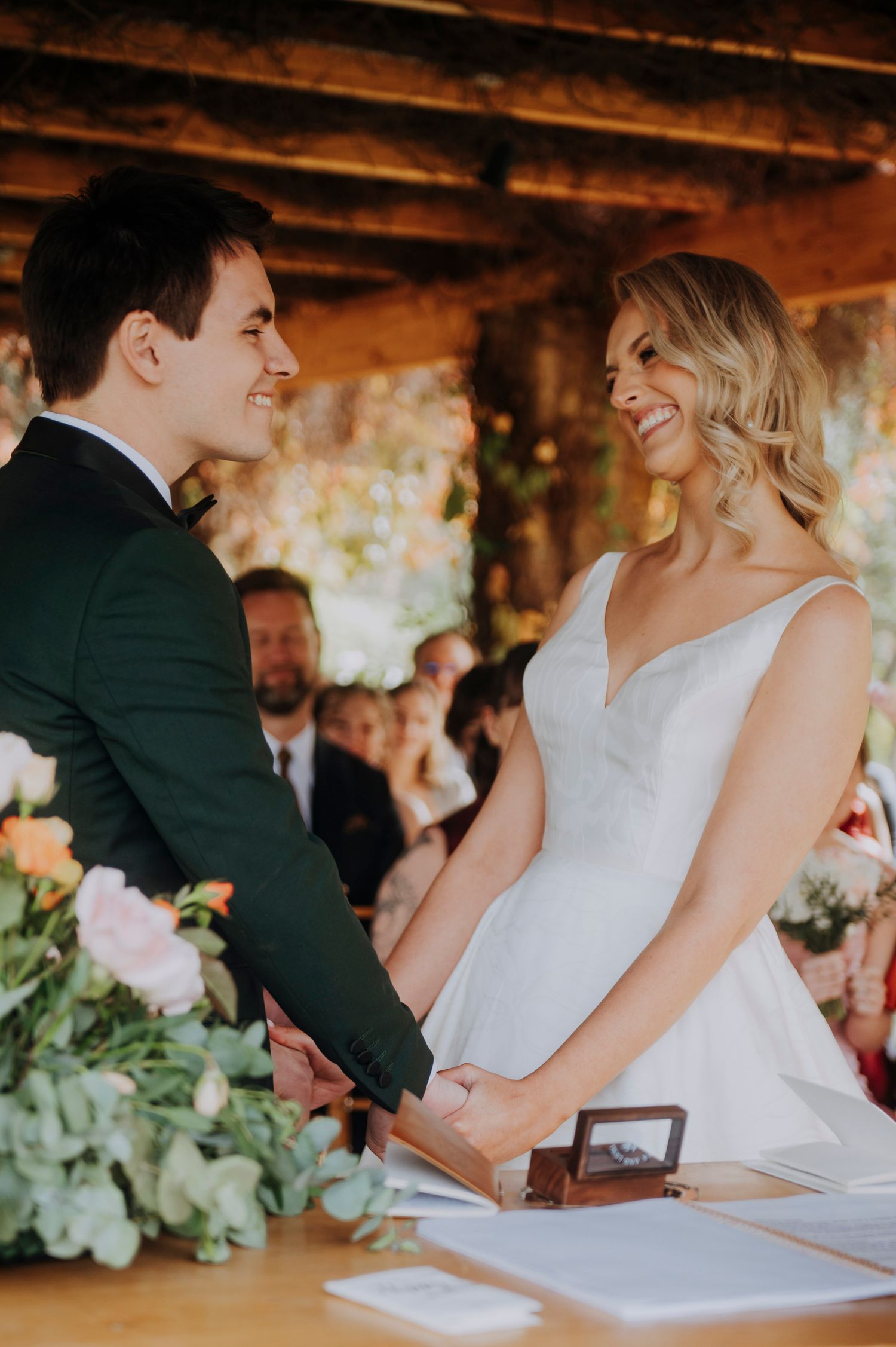 Smiling bride and groom hold hands at wedding altar, bride in white gown, groom in dark suit, floral arrangements nearby.