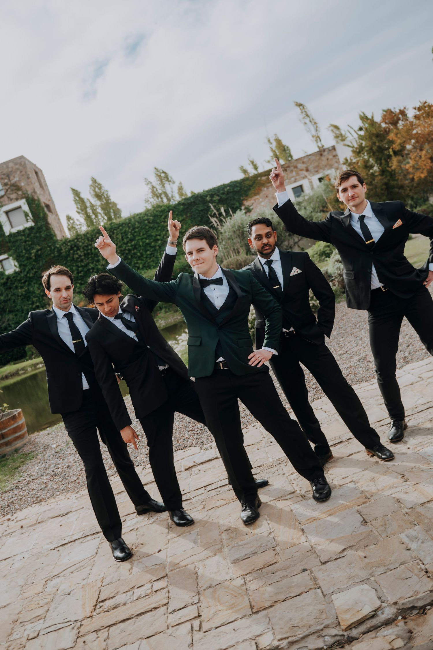 Five groomsmen in black tuxedos pose playfully outdoors, raising their arms in celebration at a wedding.