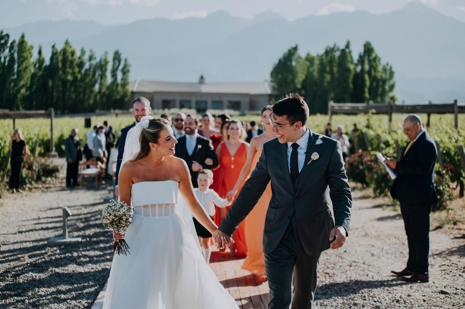 Newlyweds holding hands and smiling as they walk past guests at an outdoor vineyard wedding.