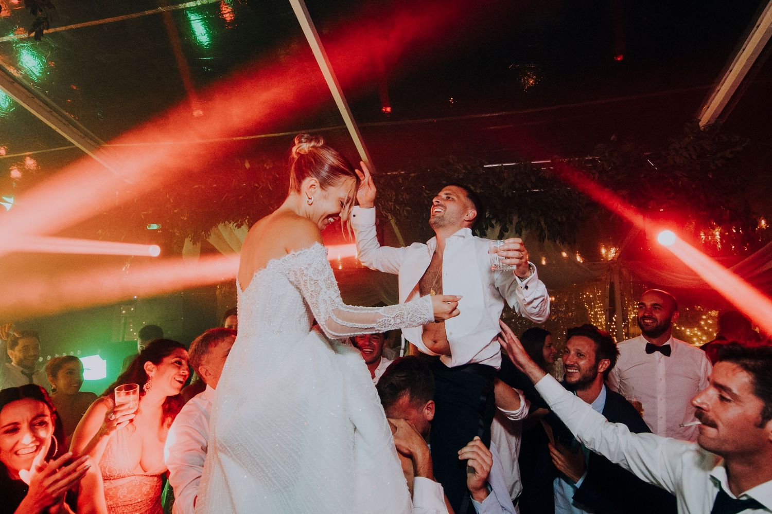 Bride and groom lifted by guests on a vibrant wedding dance floor with red laser lights.