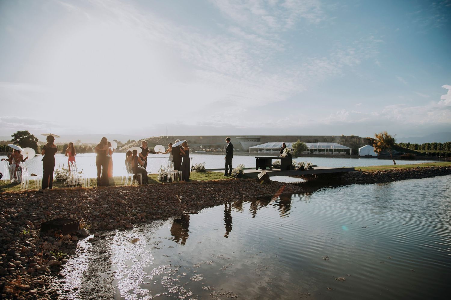 Group of people gathered by a tranquil lakeside at golden hour, with reflections on calm water and a bridge in the background.