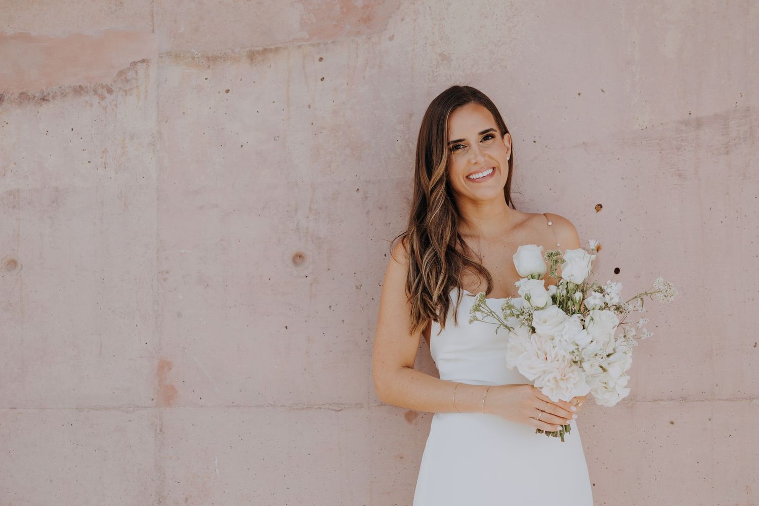 Smiling bride in strapless white gown holding white and blush bouquet against a pink stone wall.