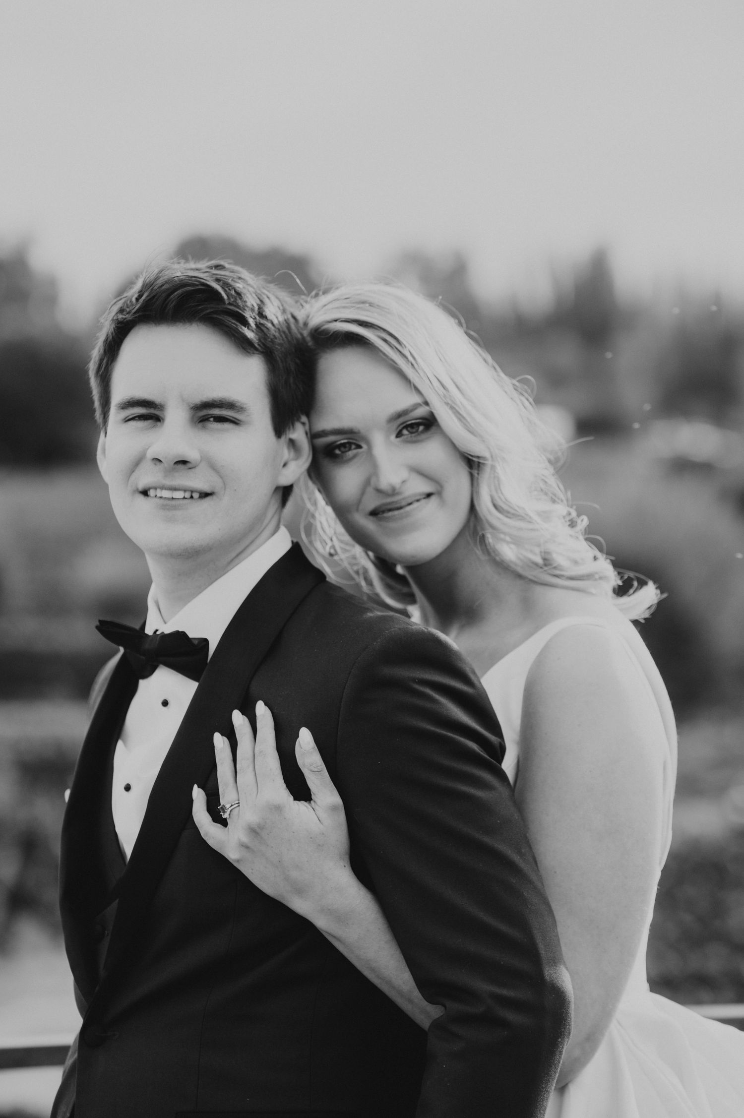 Black and white wedding portrait of smiling groom in tuxedo with bride in white dress leaning on his shoulder.