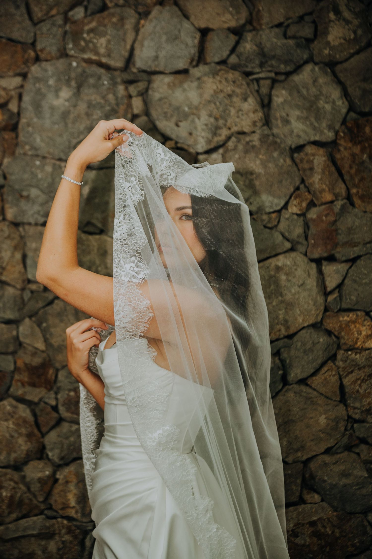 Beautiful bride in white lace wedding dress lifting her veil against a stone wall.