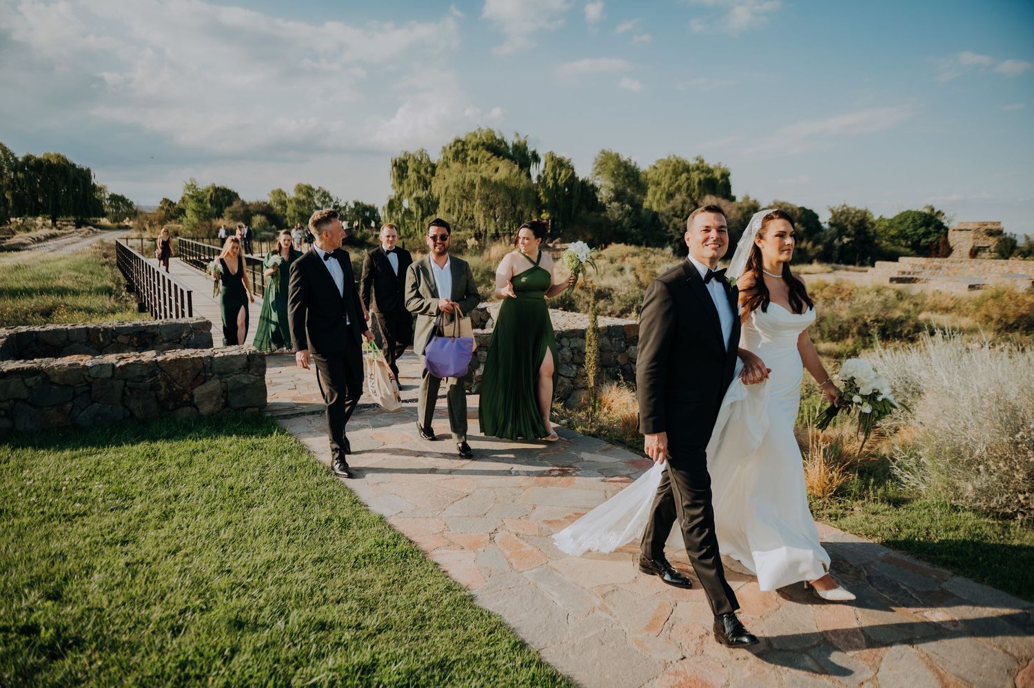 Wedding party in green dresses and black tuxedos walk along outdoor path on sunny day.