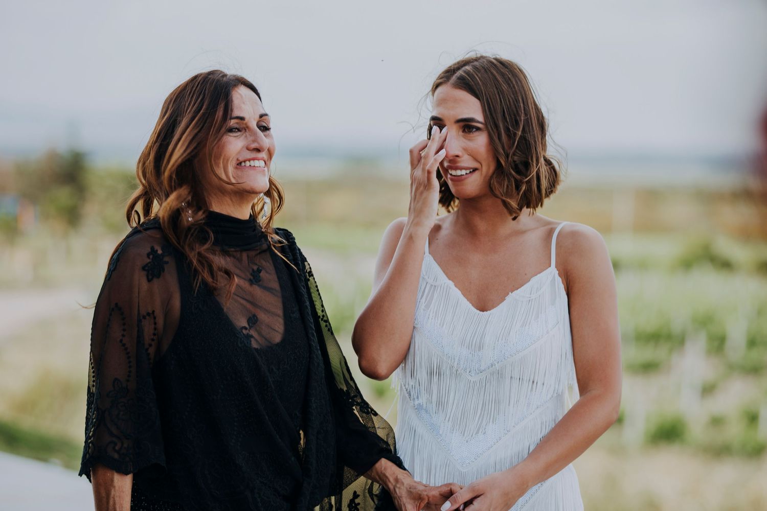 Two women smile emotionally outdoors, one in a white dress wiping tears, likely at a wedding celebration.