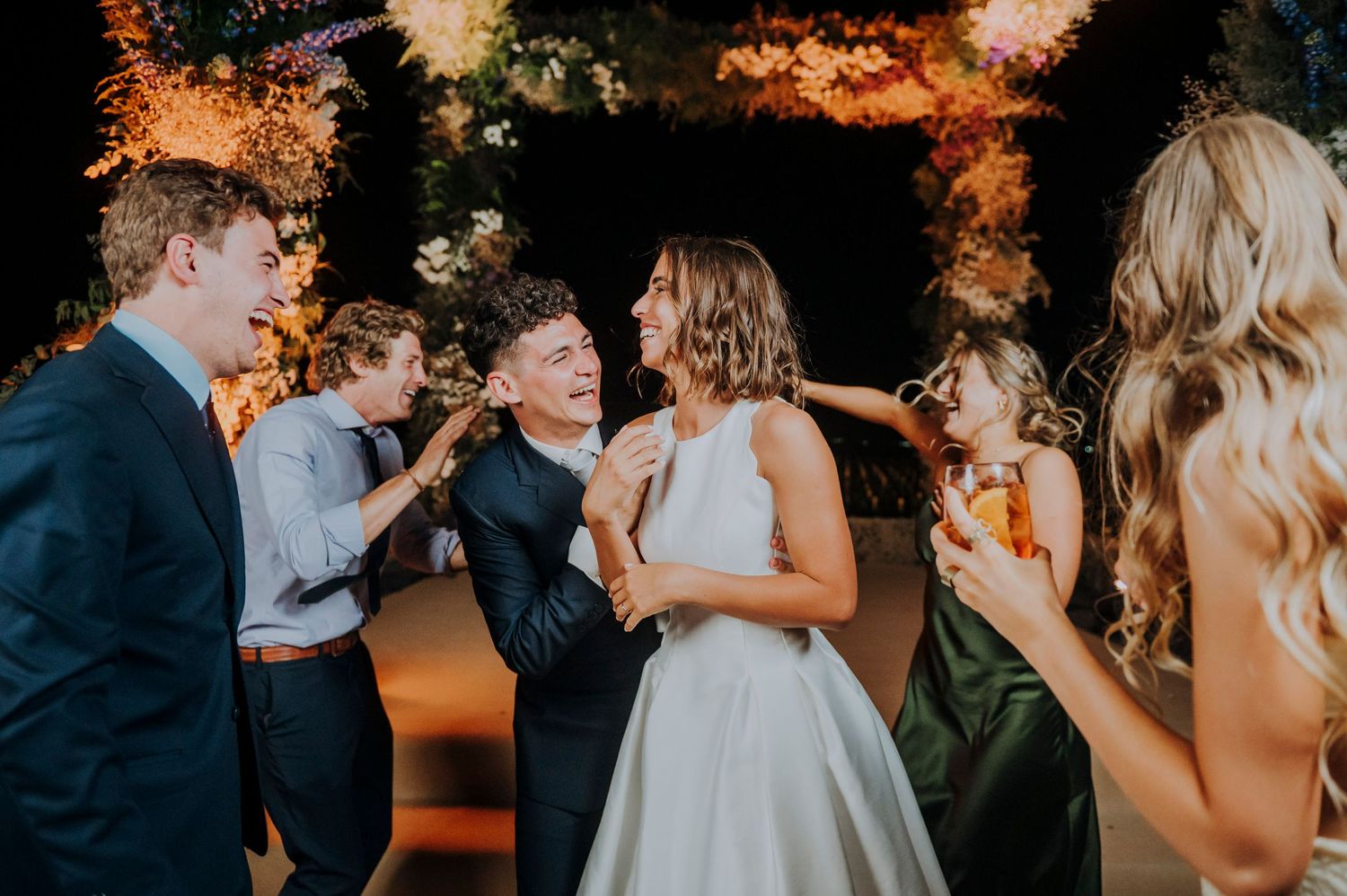 Bride laughs with guests during a lively outdoor wedding reception celebration at night.