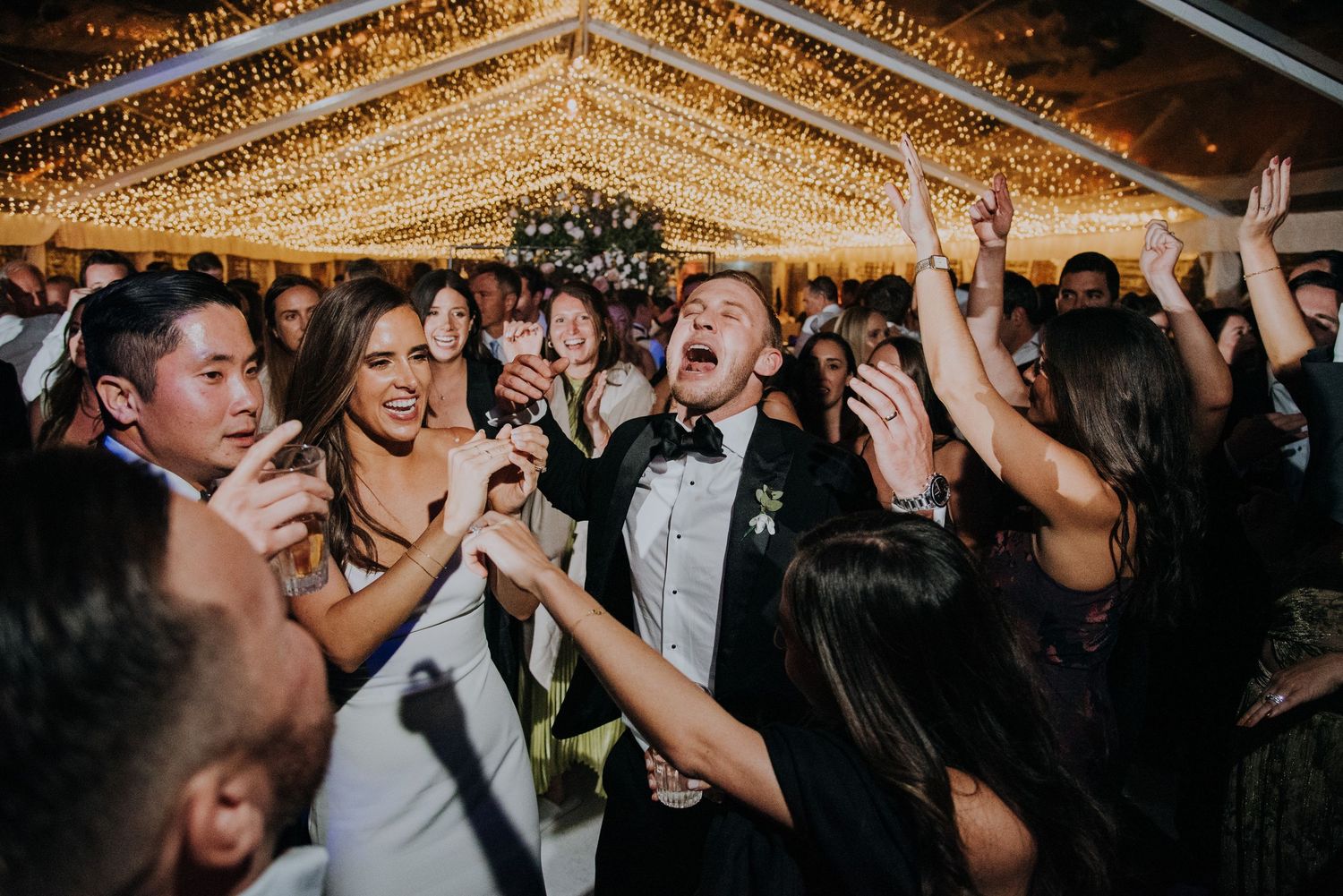 Joyful wedding reception dance floor with guests celebrating under warm fairy lights in a tented venue.