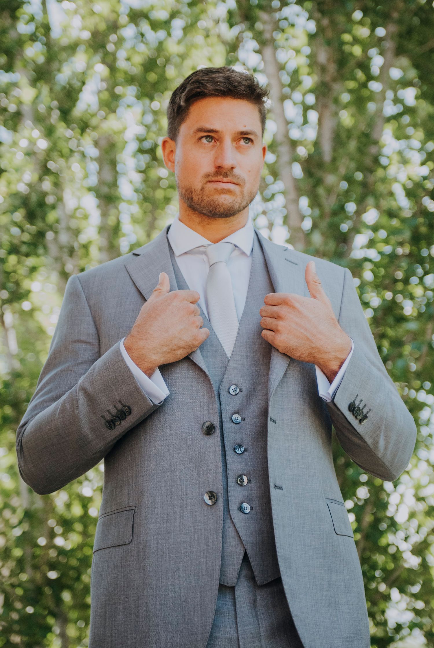 Confident groom adjusting his grey three-piece suit jacket while standing outdoors among green trees in Mendoza