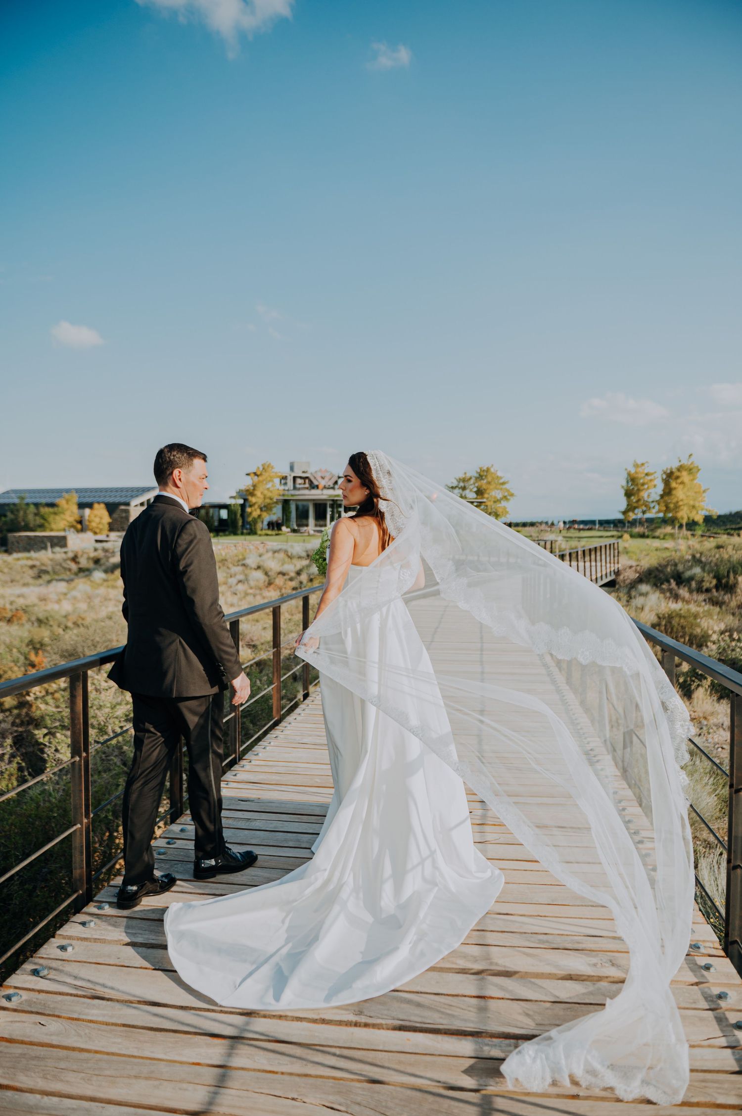 Bride in flowing white gown with long veil and groom on wooden bridge with autumn landscape backdrop Couple portrait in Huentala Mendoza.