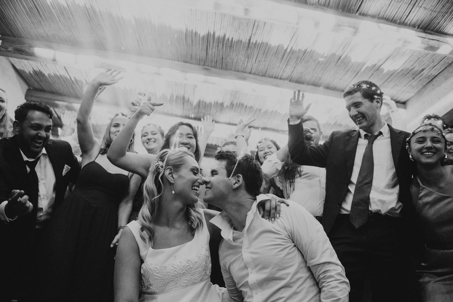 Bride and groom share a romantic kiss surrounded by cheering wedding guests in joyful black and white photo.