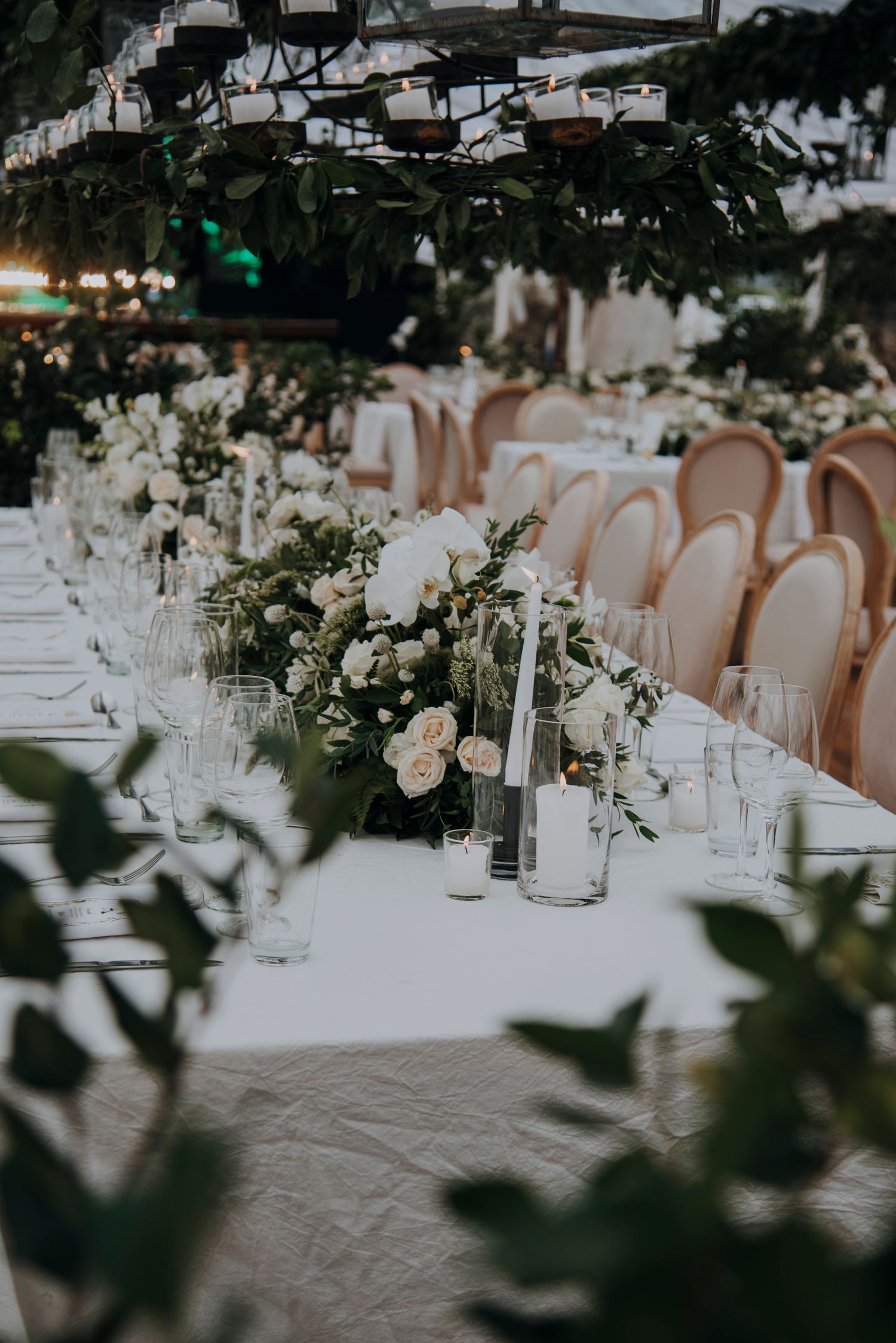 Elegant wedding reception table with white floral centerpieces, greenery, candles, and blush chairs.