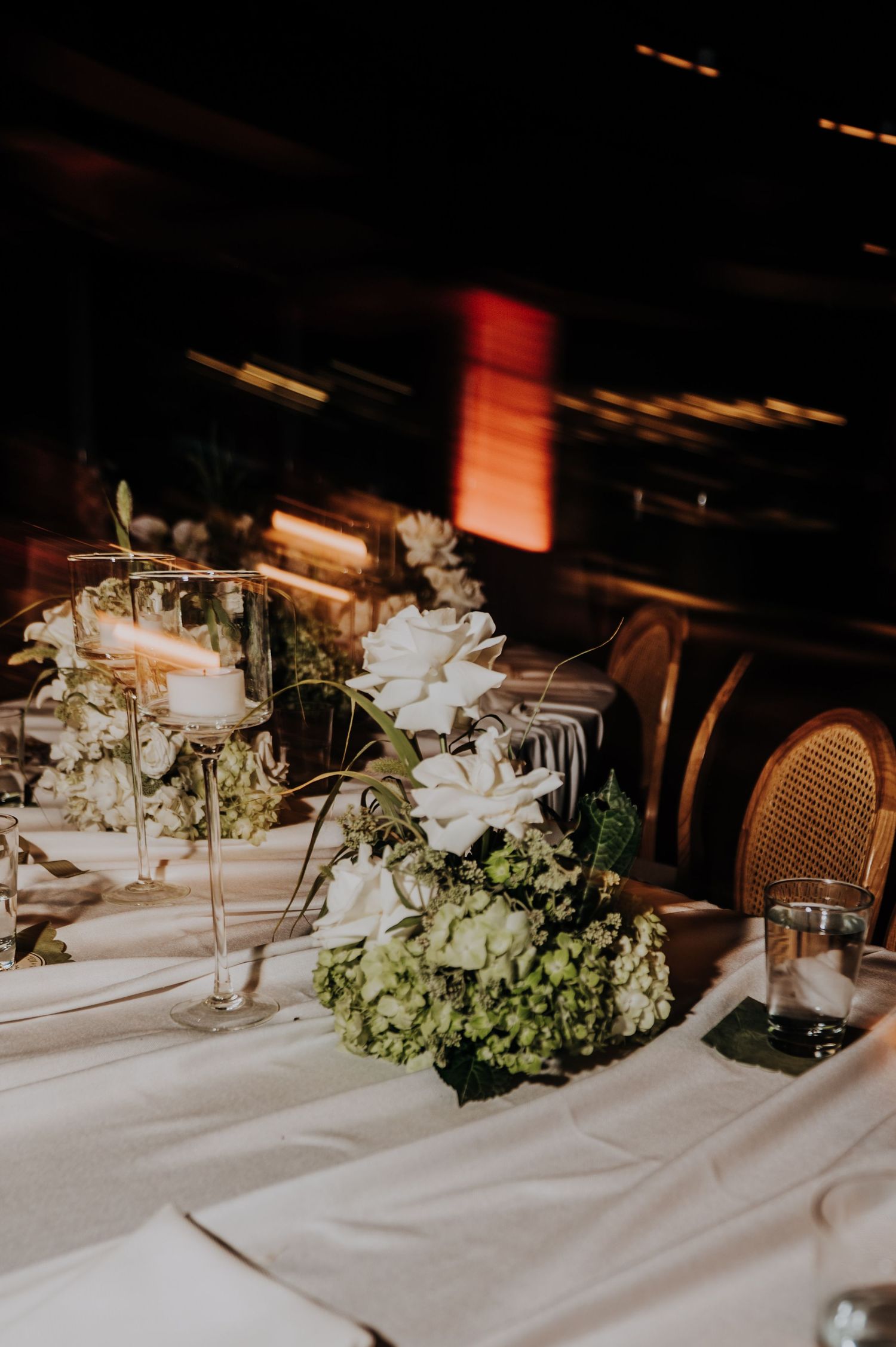 Elegant wedding reception table with white and green floral centerpieces and candlelight in dimly lit venue.