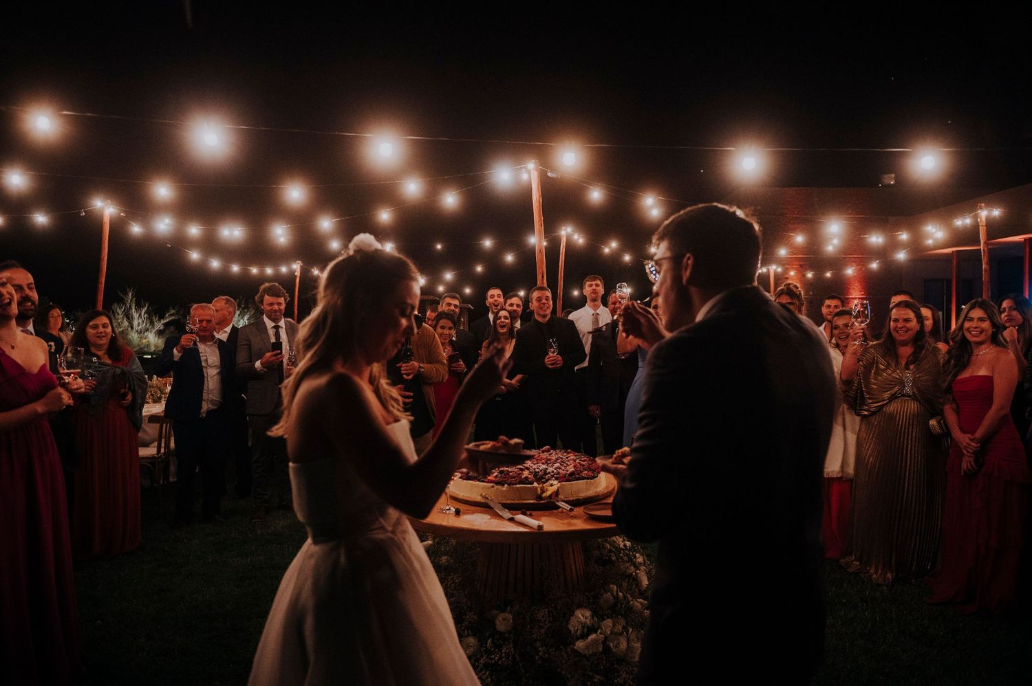 Bride and groom cut wedding cake surrounded by guests holding sparklers at night.