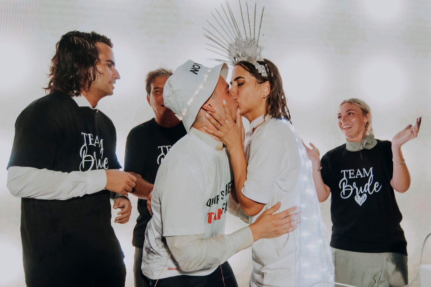 Bride wearing a silver crown kisses partner while Team Bride group celebrates at a bachelorette party.