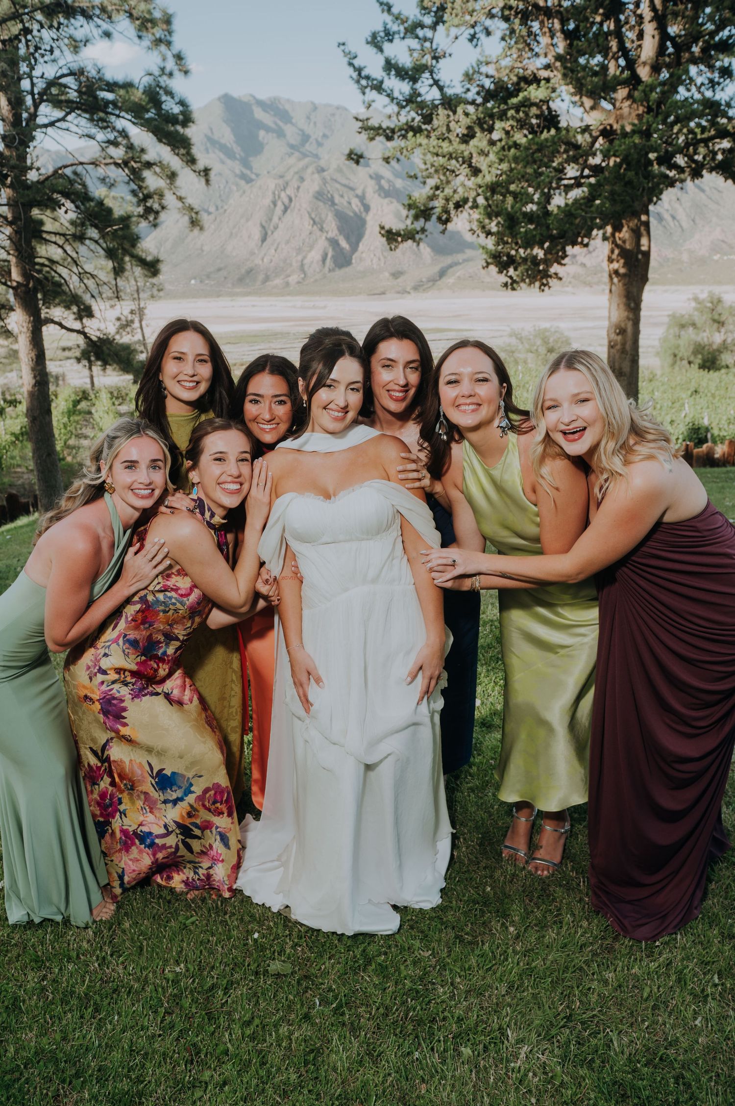 Bride surrounded by smiling bridesmaids in colorful dresses with mountain backdrop.