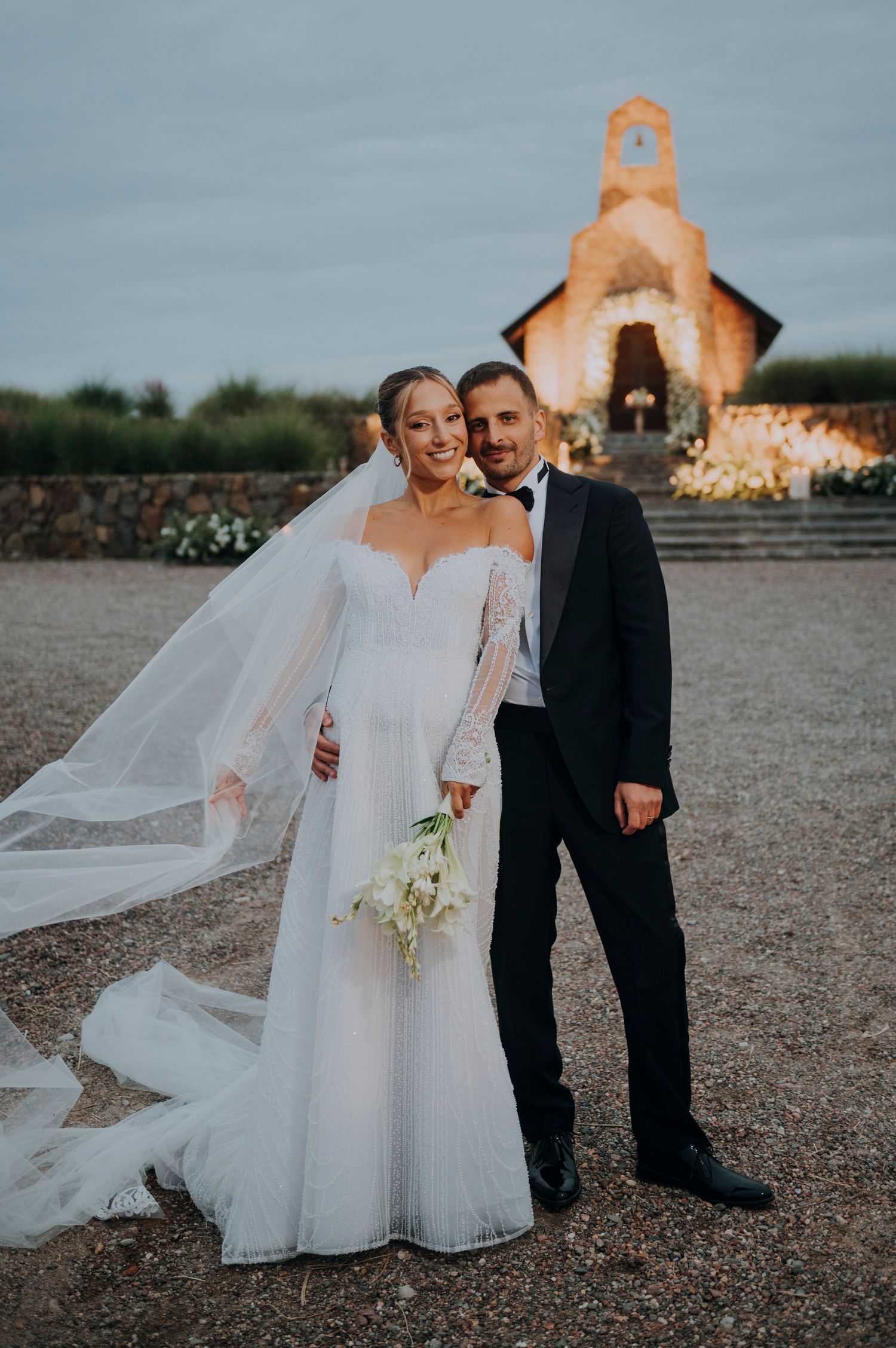 Bride in white gown with veil and groom in tuxedo posing in front of a lit chapel at dusk.