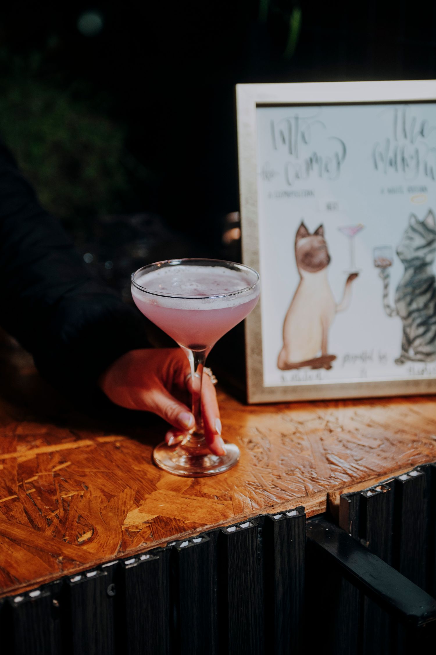 A hand holds a pink cocktail in a coupe glass beside a framed cat illustration at a wedding bar.