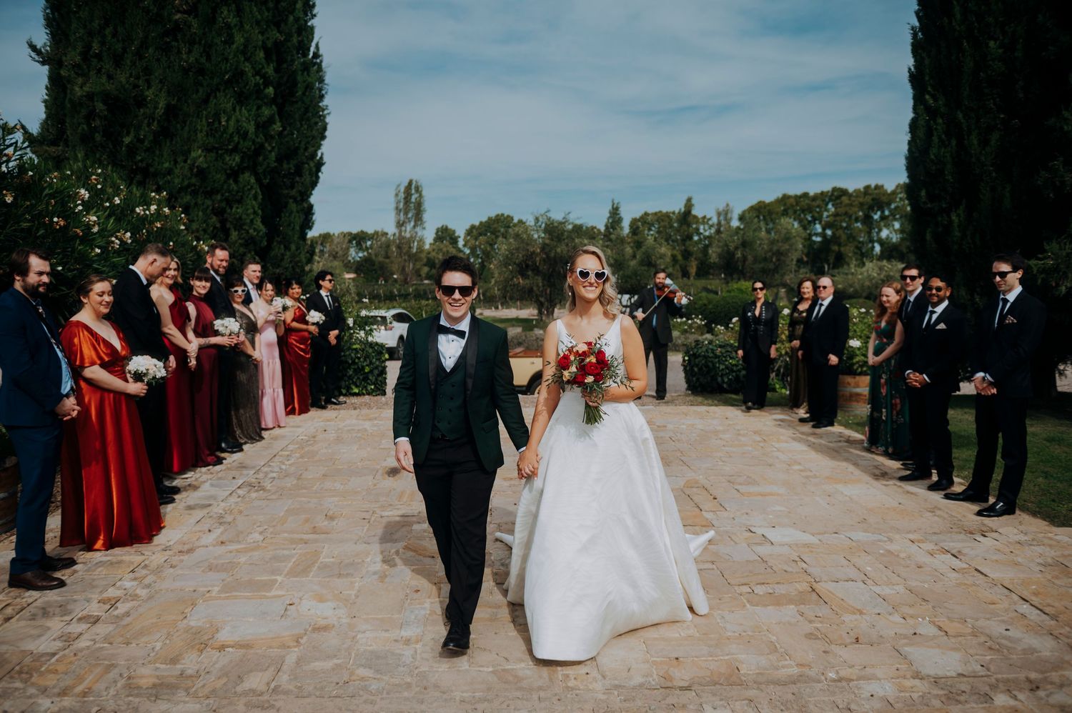 Bride and groom walk hand-in-hand down stone aisle lined with wedding guests at outdoor garden ceremony.