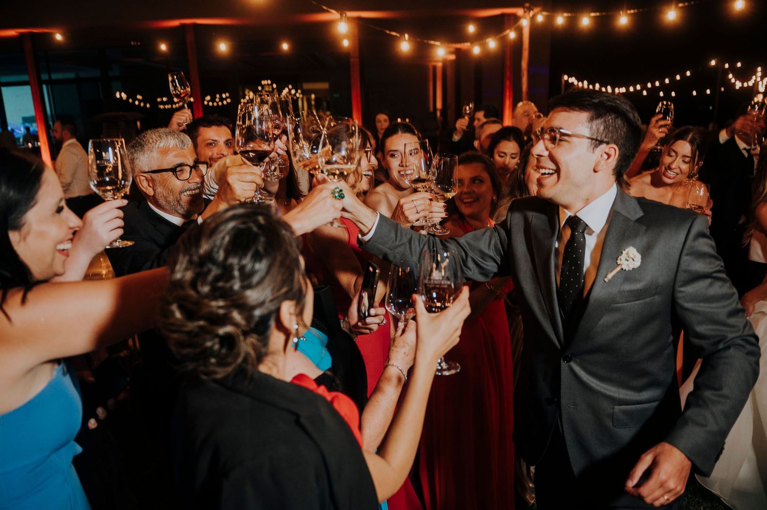 Groom toasts with guests at a festive wedding reception under warm string lights.
