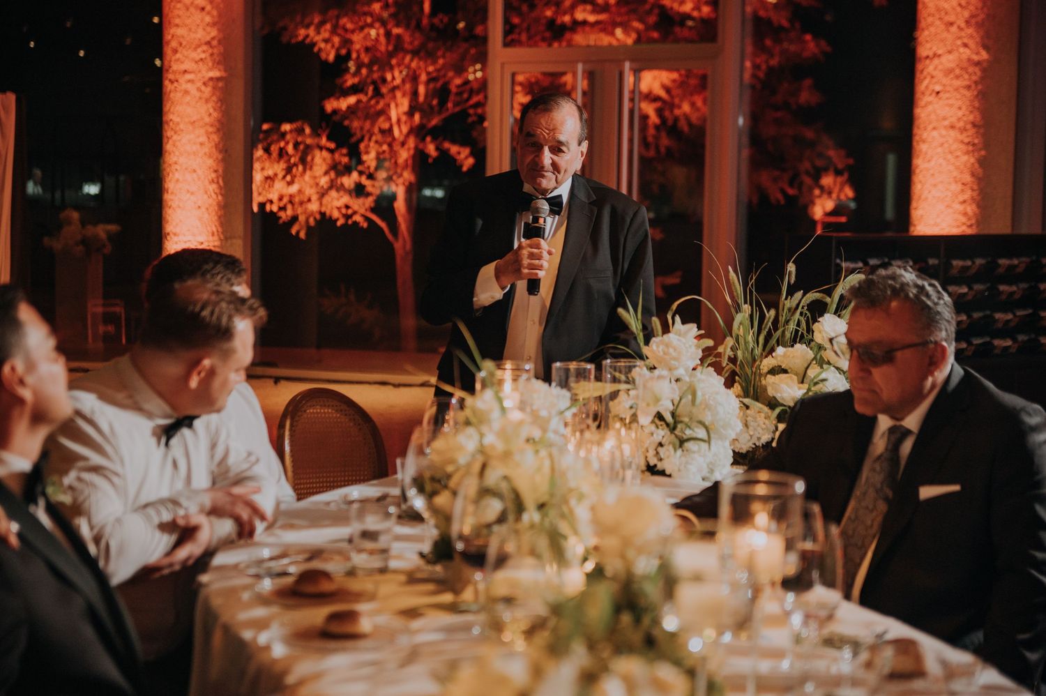 Wedding guest delivering speech at elegantly decorated reception dinner table with floral centerpieces.