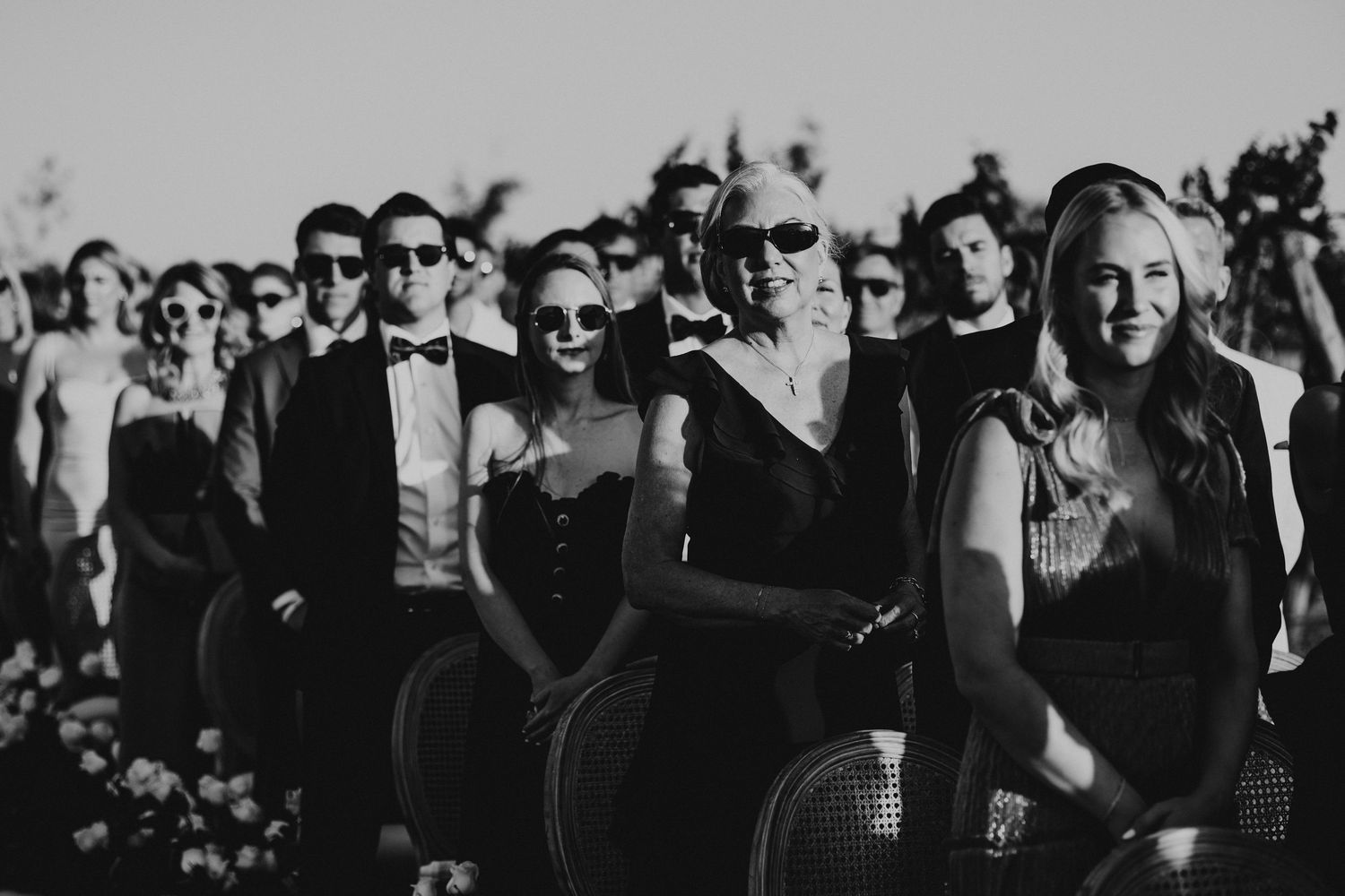 Black and white photo of wedding guests seated and standing at an outdoor ceremony in sunglasses.