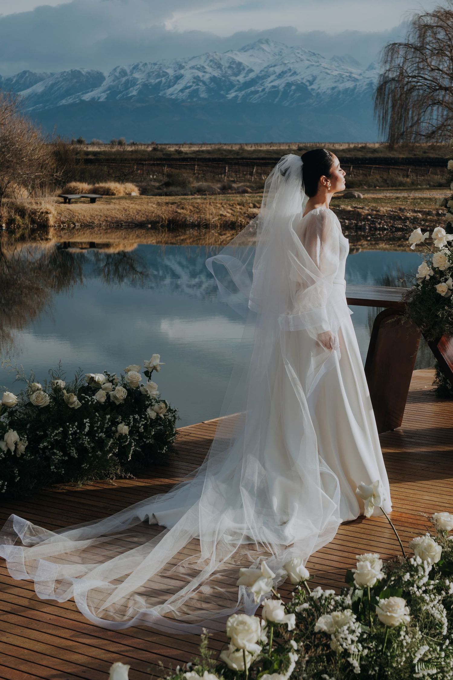 Elegant bride in cathedral veil standing on wooden dock with snowy mountain backdrop in huentala mendoza